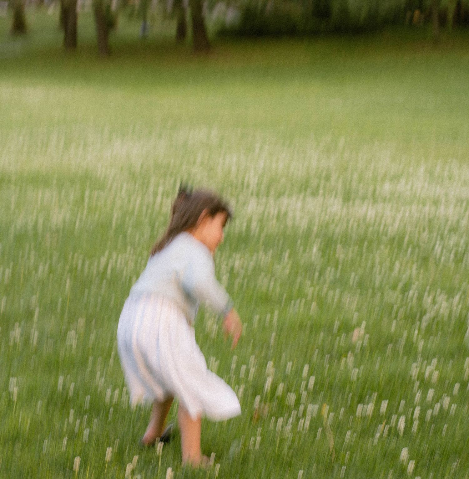 A young girl in a white dress plays in a field of tall grass and wildflowers, captured in soft motion blur.