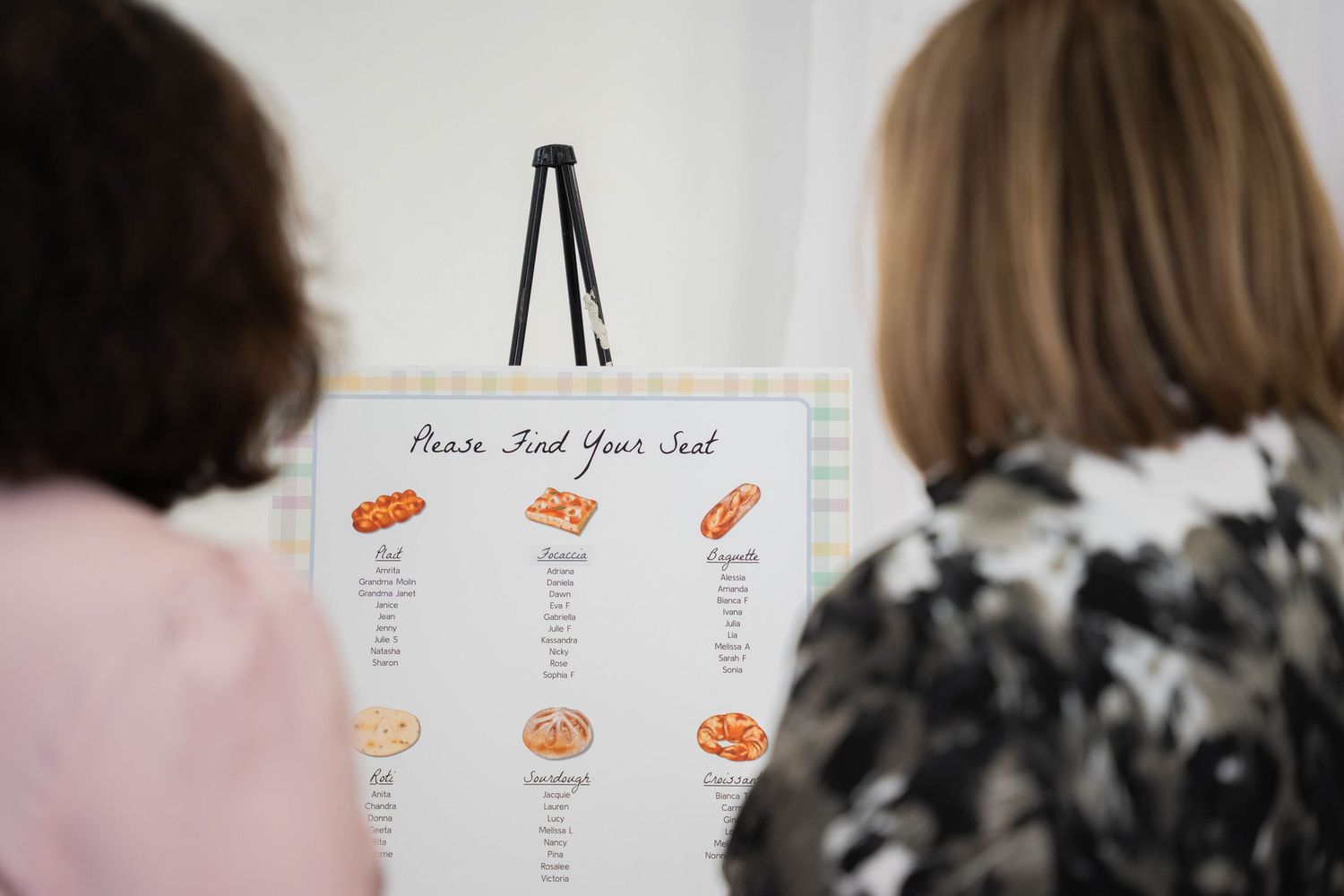 Two guests look at a Please Find Your Seat chart decorated with bread illustrations at a baby shower.