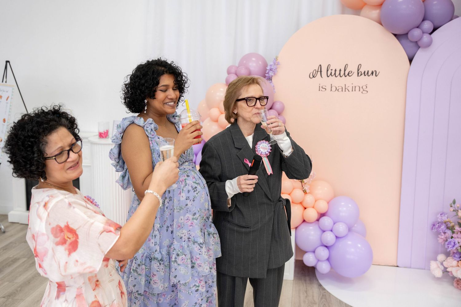 Three guests hold drinks and smile at a baby shower in front of a peach arch sign reading a little bun is baking.