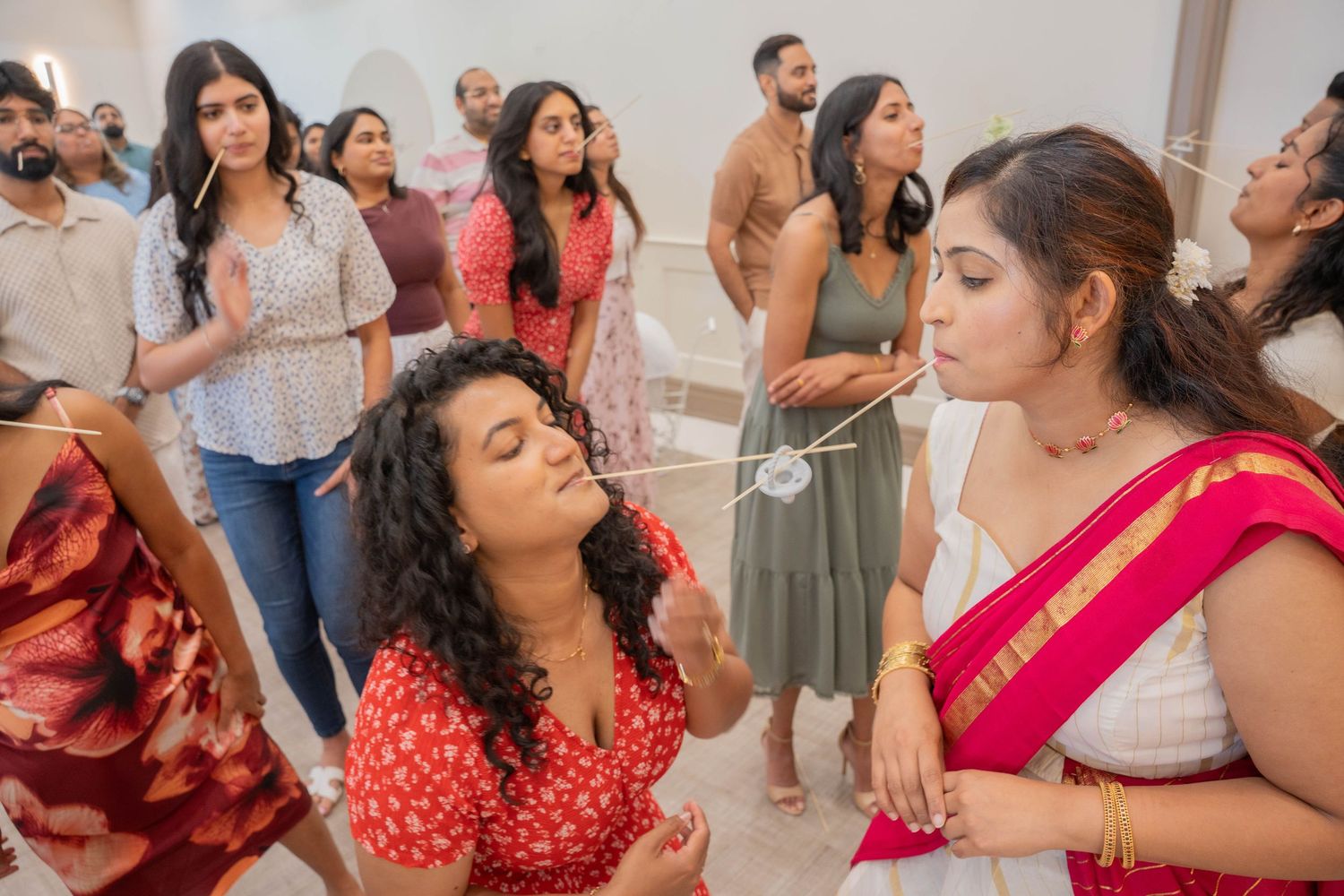 Guests laugh and cheer as two women compete in a baby shower game involving strings held in their mouths.