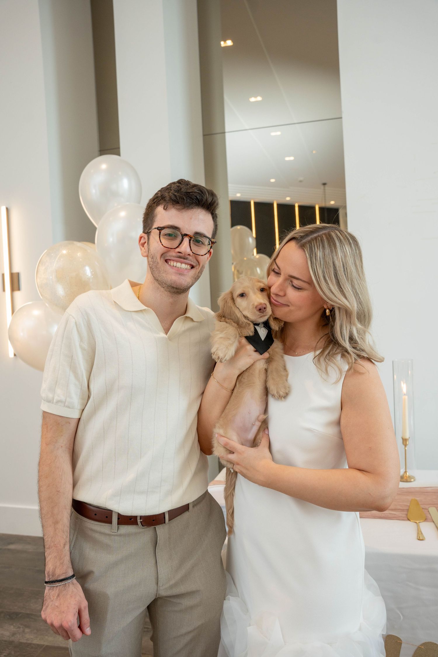Smiling couple at an engagement celebration hold a fluffy puppy in a bow tie in front of white balloons.