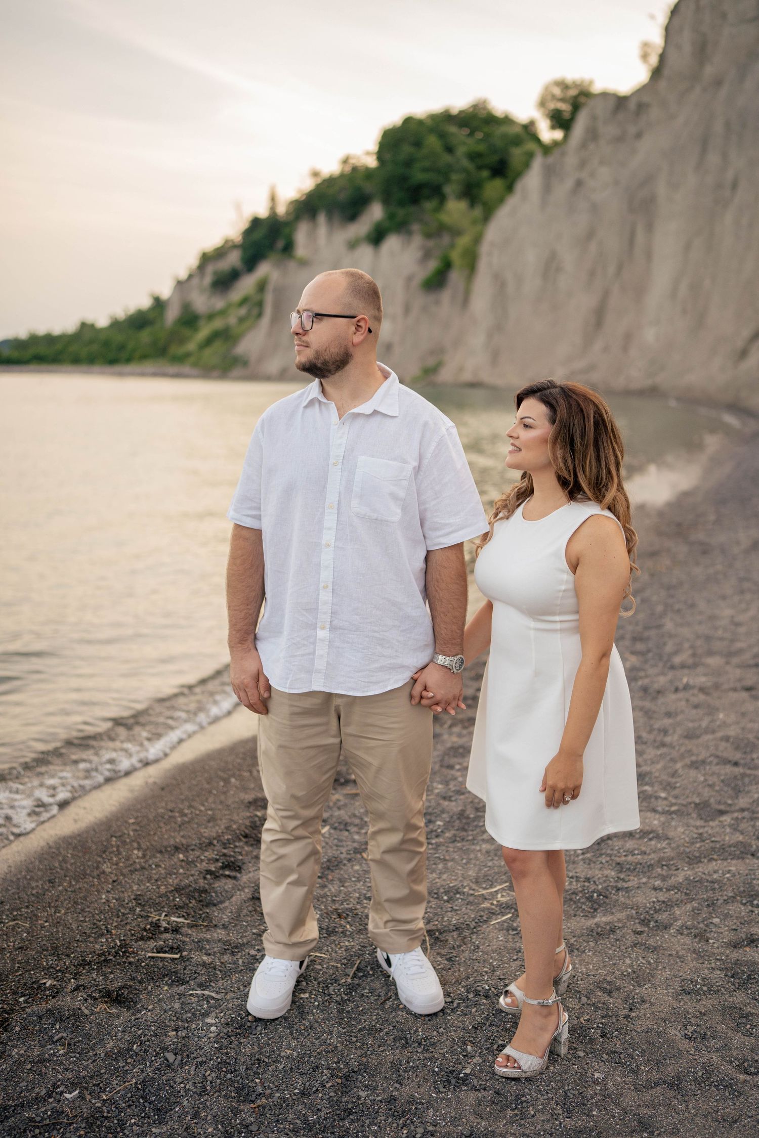 Smiling couple in white outfits holding hands on a pebble beach beside calm water and towering lakeside cliffs.