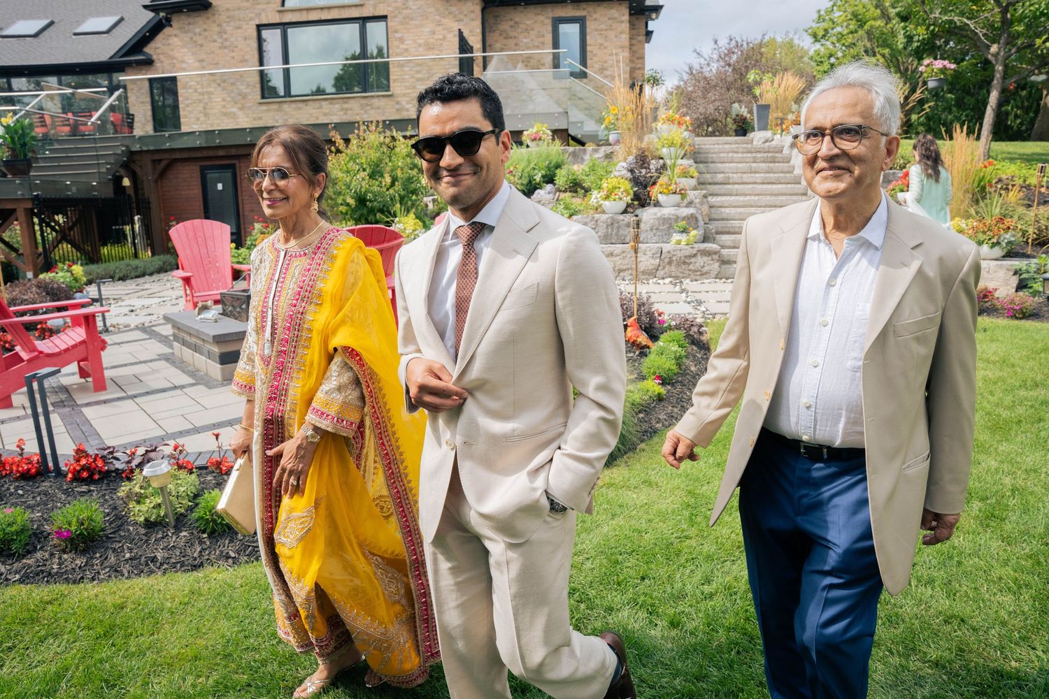 A smiling groom in sunglasses and a tan suit strolls across a lawn with his mother in a yellow shawl and his father beside him.