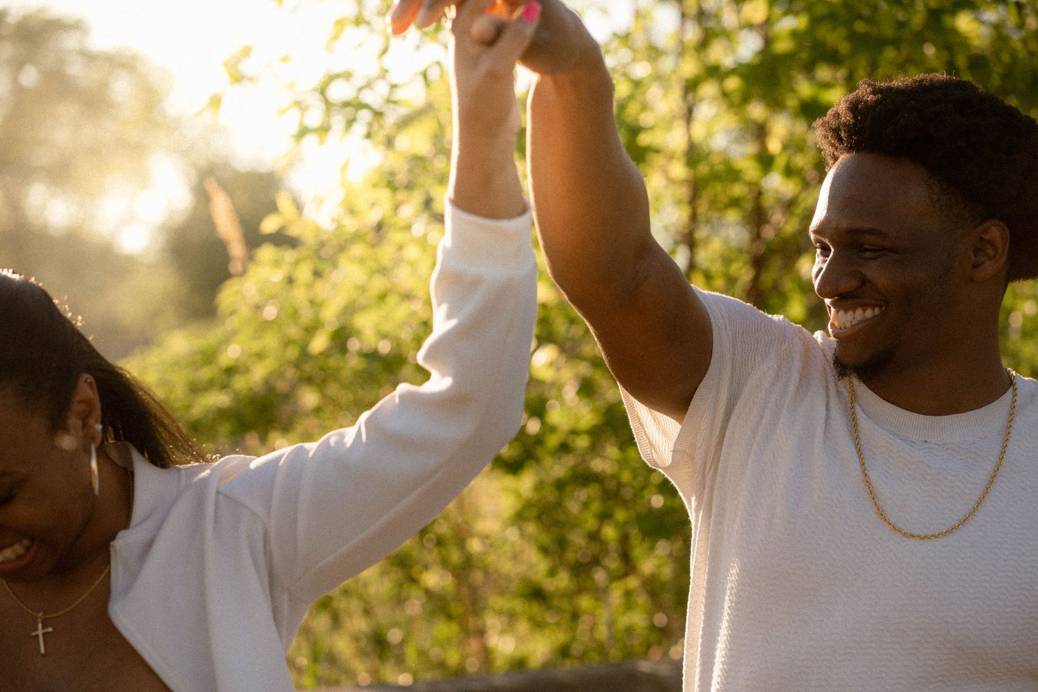 A smiling man and woman in white shirts raise their joined hands in celebration outdoors in warm golden sunlight.