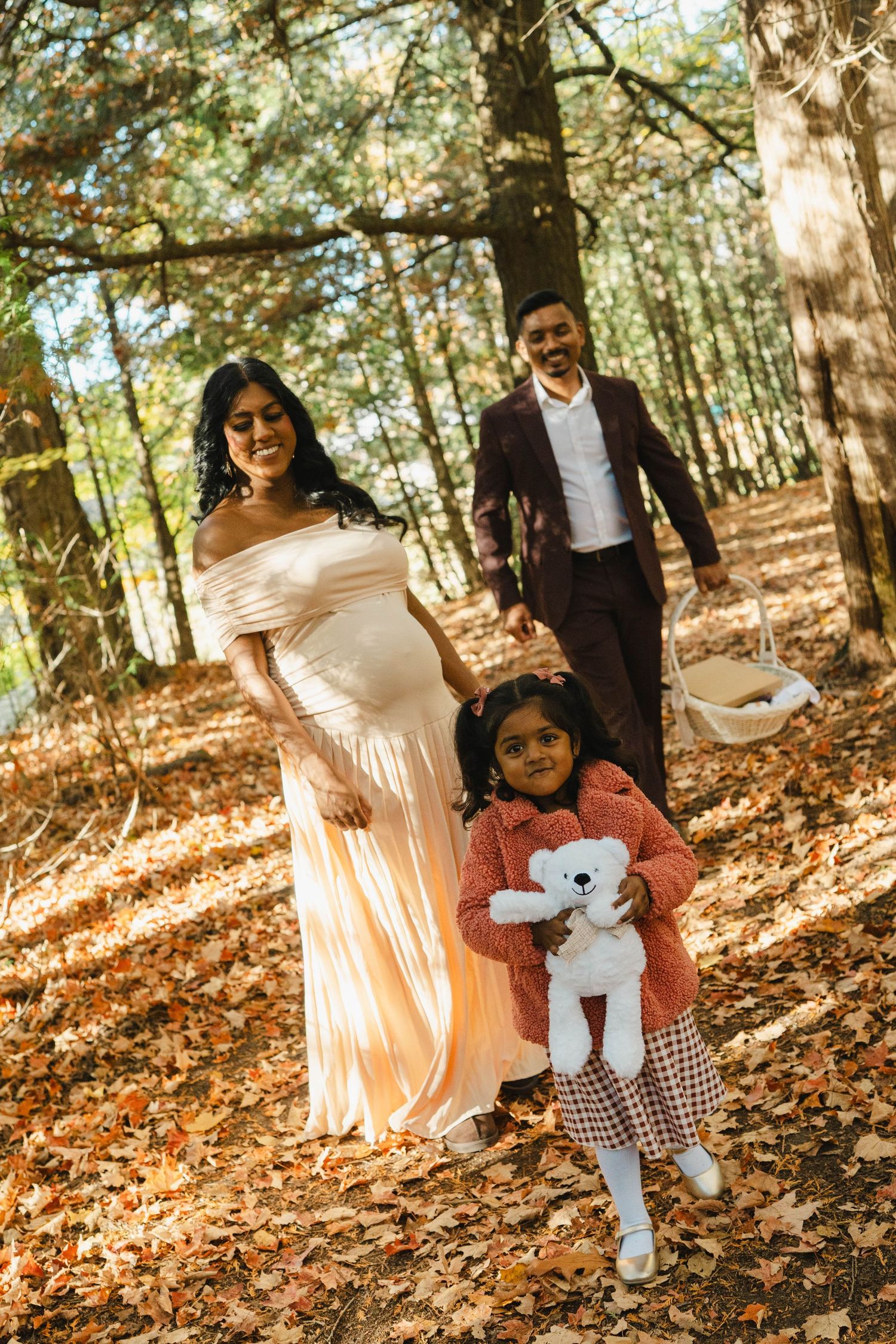 Expecting mother in a peach gown and father in a maroon suit walk through autumn woods with their young daughter holding a teddy bear.