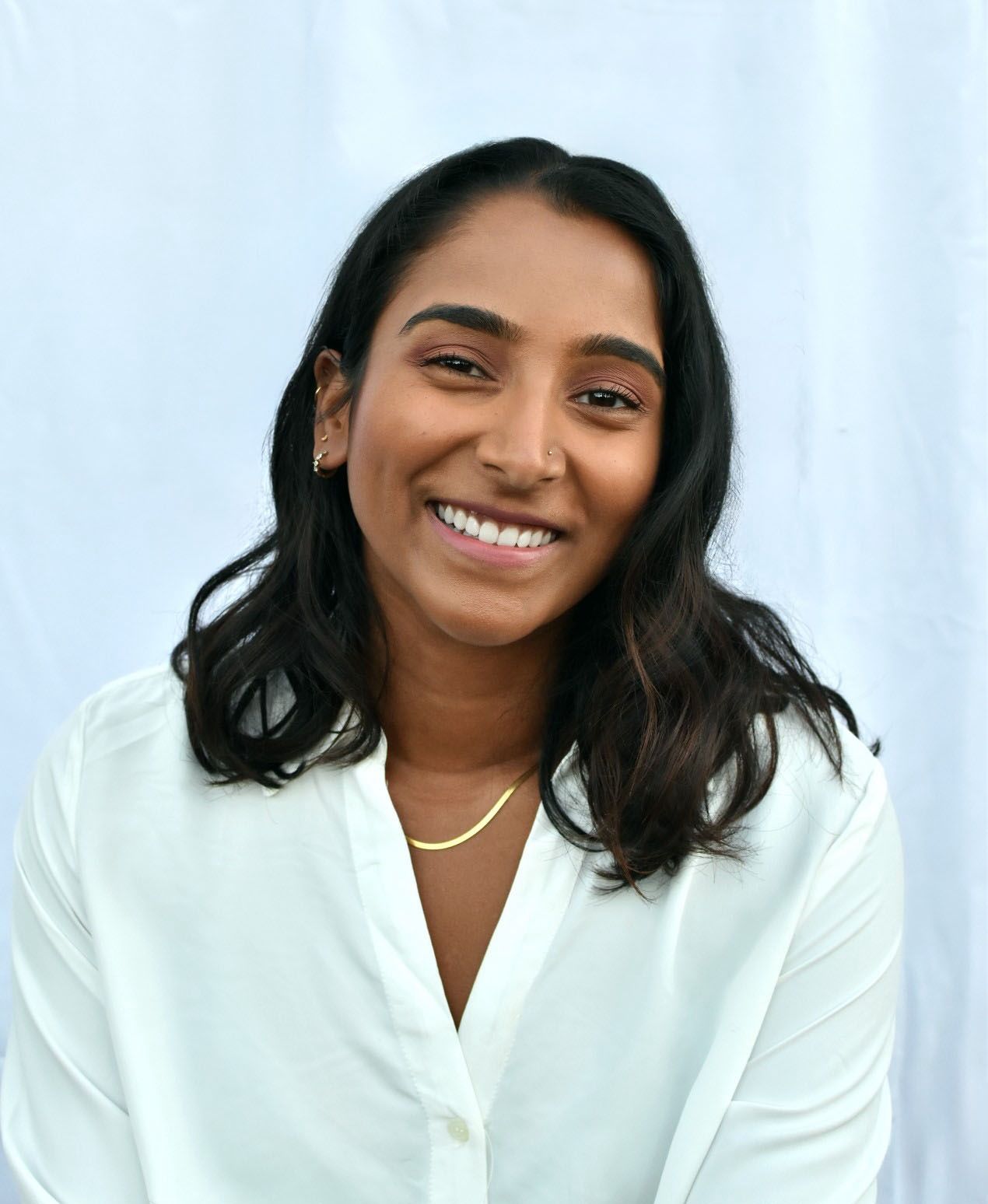 A smiling woman with shoulder-length dark hair wears a white button-up blouse and gold necklace against a white backdrop.