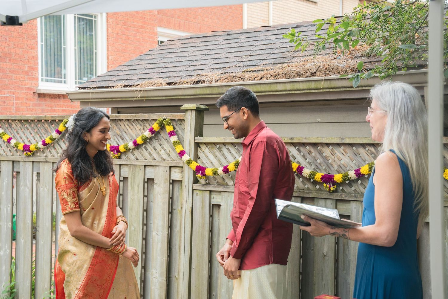 A bride and groom in traditional Indian attire stand smiling in front of an officiant reading from a book at a backyard wedding.
