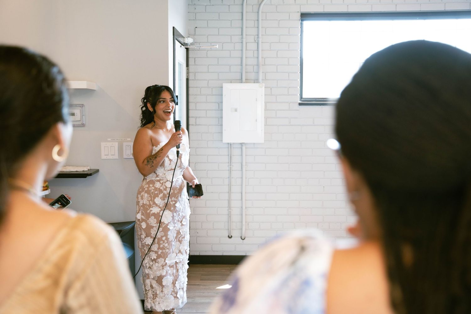 A woman in a white floral dress speaks into a microphone to seated guests during a bridal shower in a bright room.