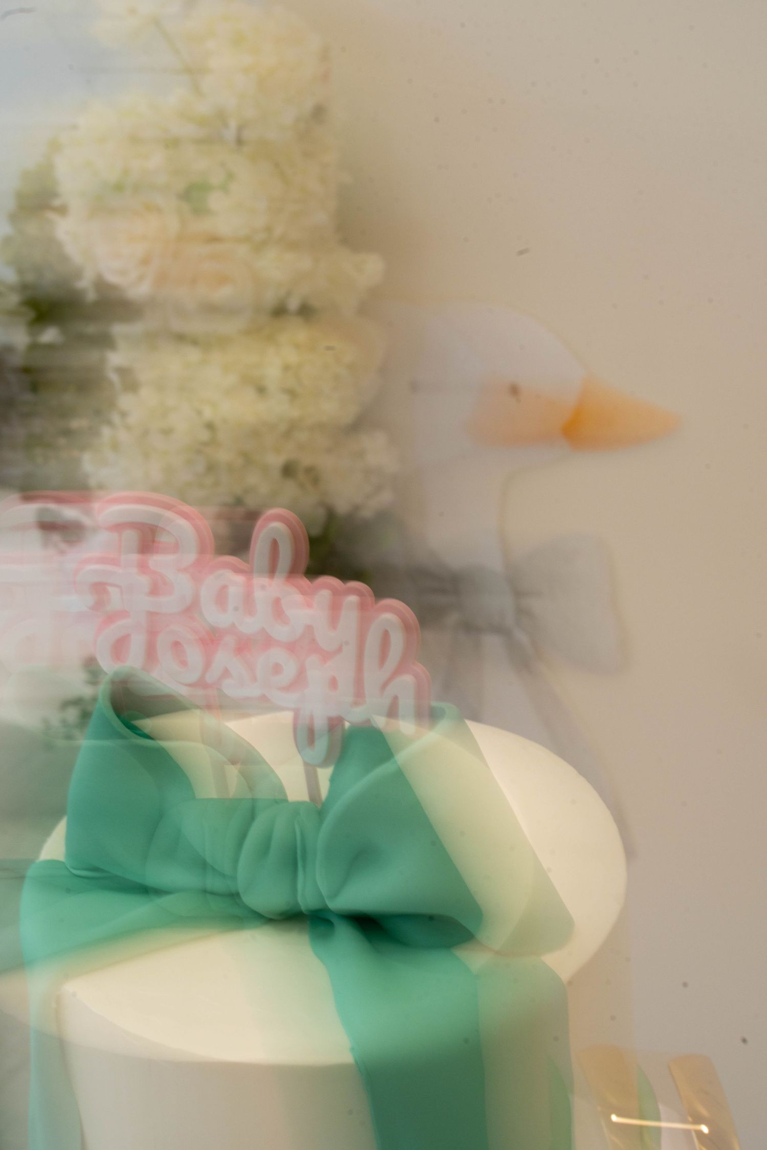A softly blurred, double-exposure baby shower cake topper with pink lettering, a teal bow, a white duck, and white blossoms.