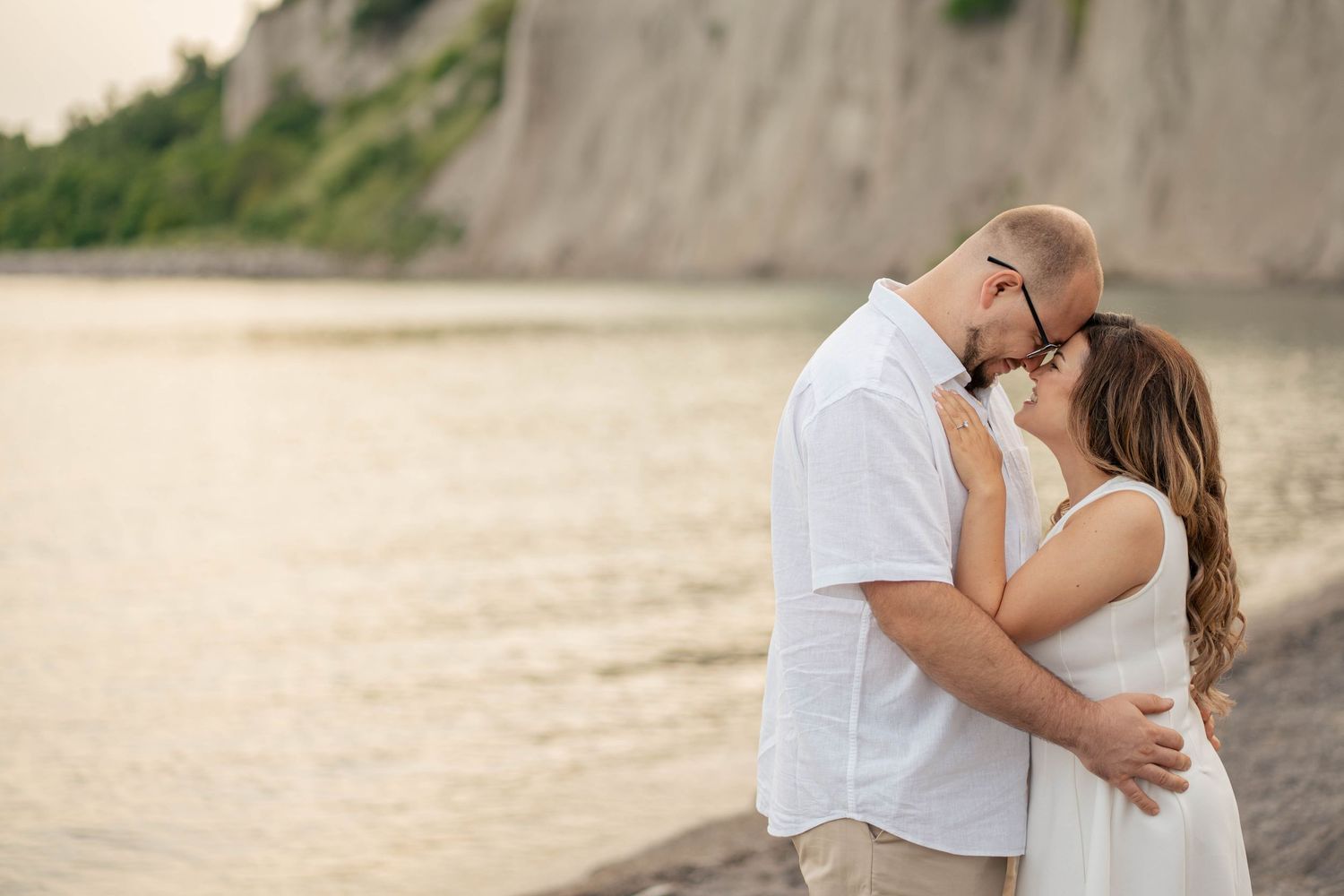 Couple in white clothing embracing close on a quiet shoreline at sunset, with soft light on the water and cliffs behind.