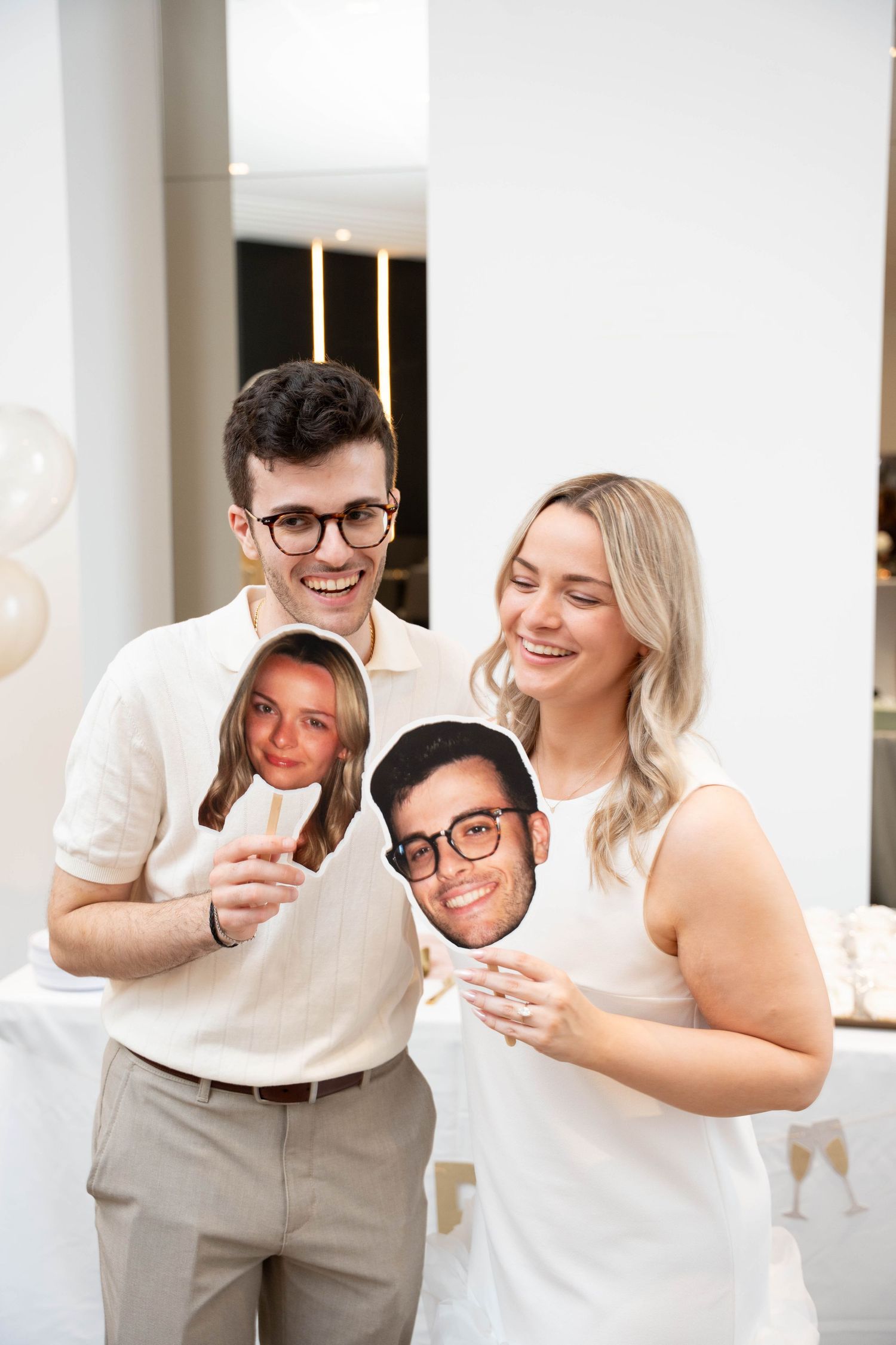 A laughing man and woman hold large cutout faces on sticks at a bright engagement party with white decor and champagne glasses.