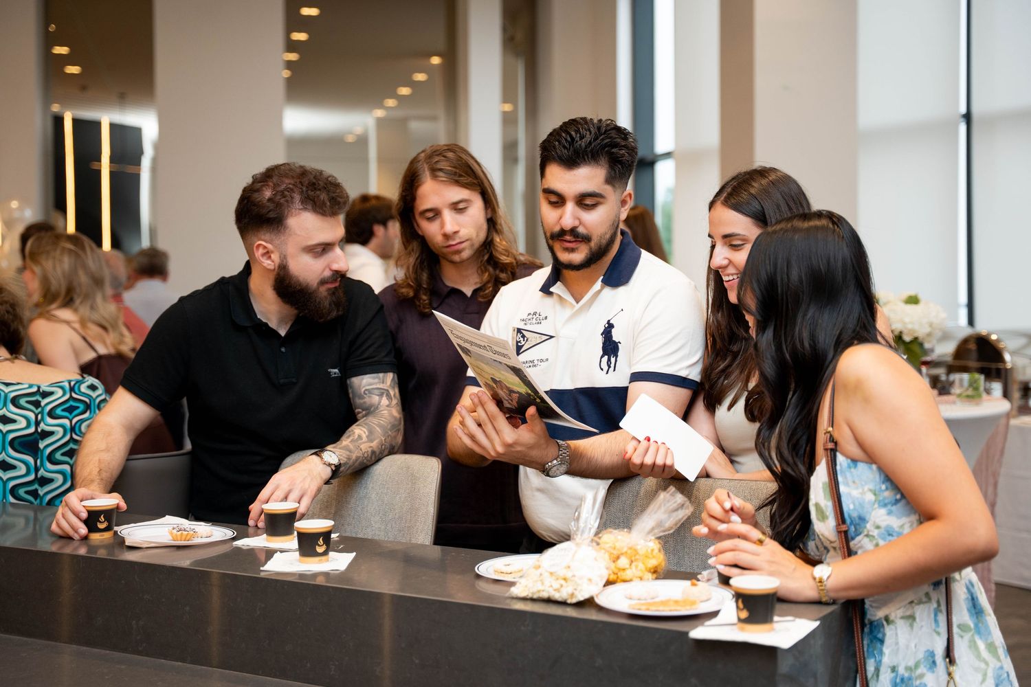 A group of friends gathers around a table with coffee and desserts, reading an engagement party newspaper together.