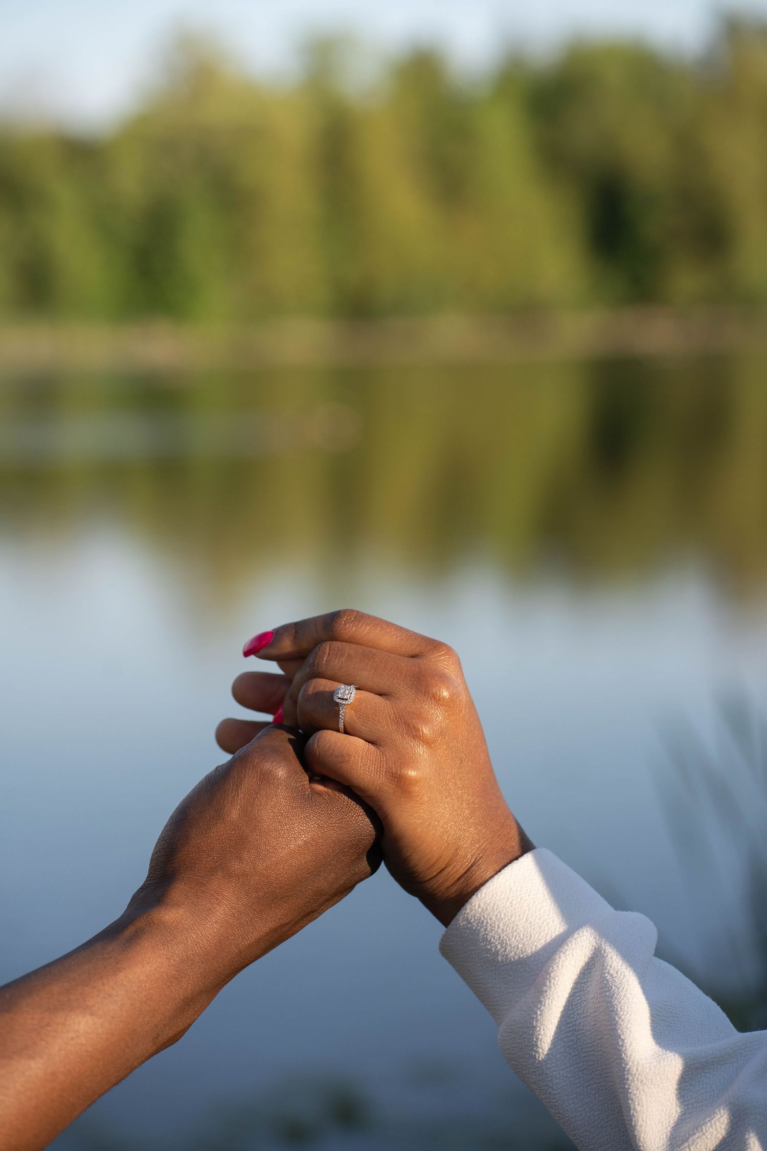 Close-up of two hands clasped together, one wearing a diamond engagement ring, with a calm lake and green trees in the background.