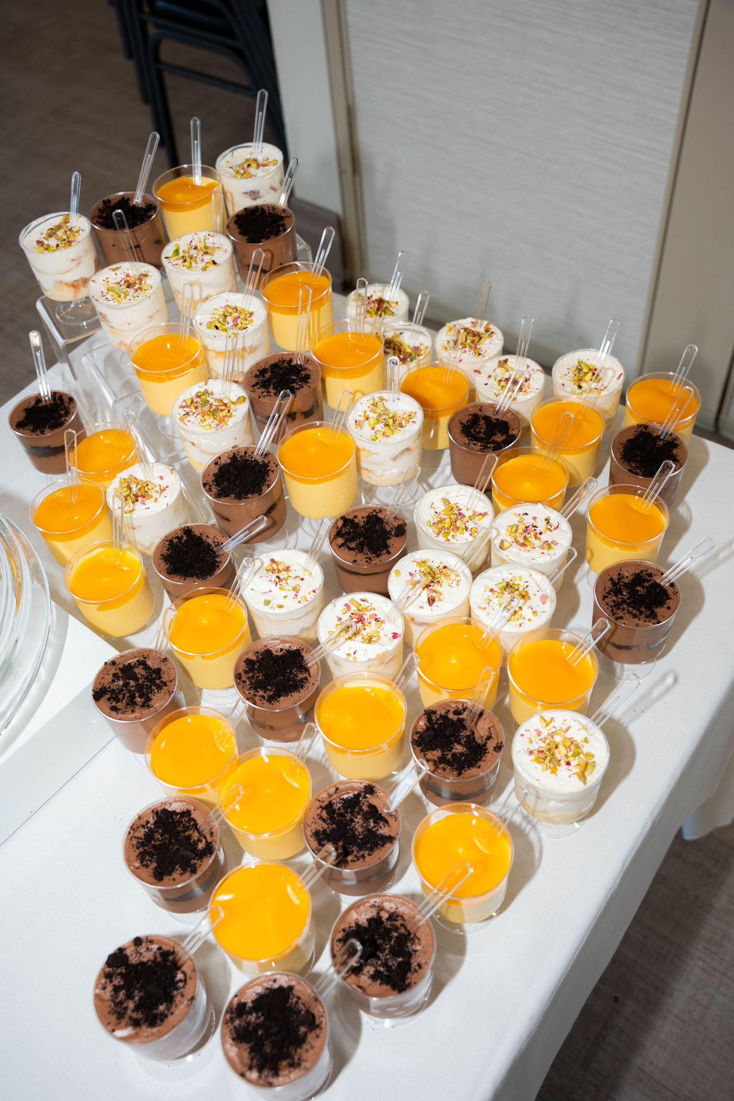 Rows of mini dessert cups with mango mousse, chocolate cookie crumble, and creamy toppings arranged on a white table.