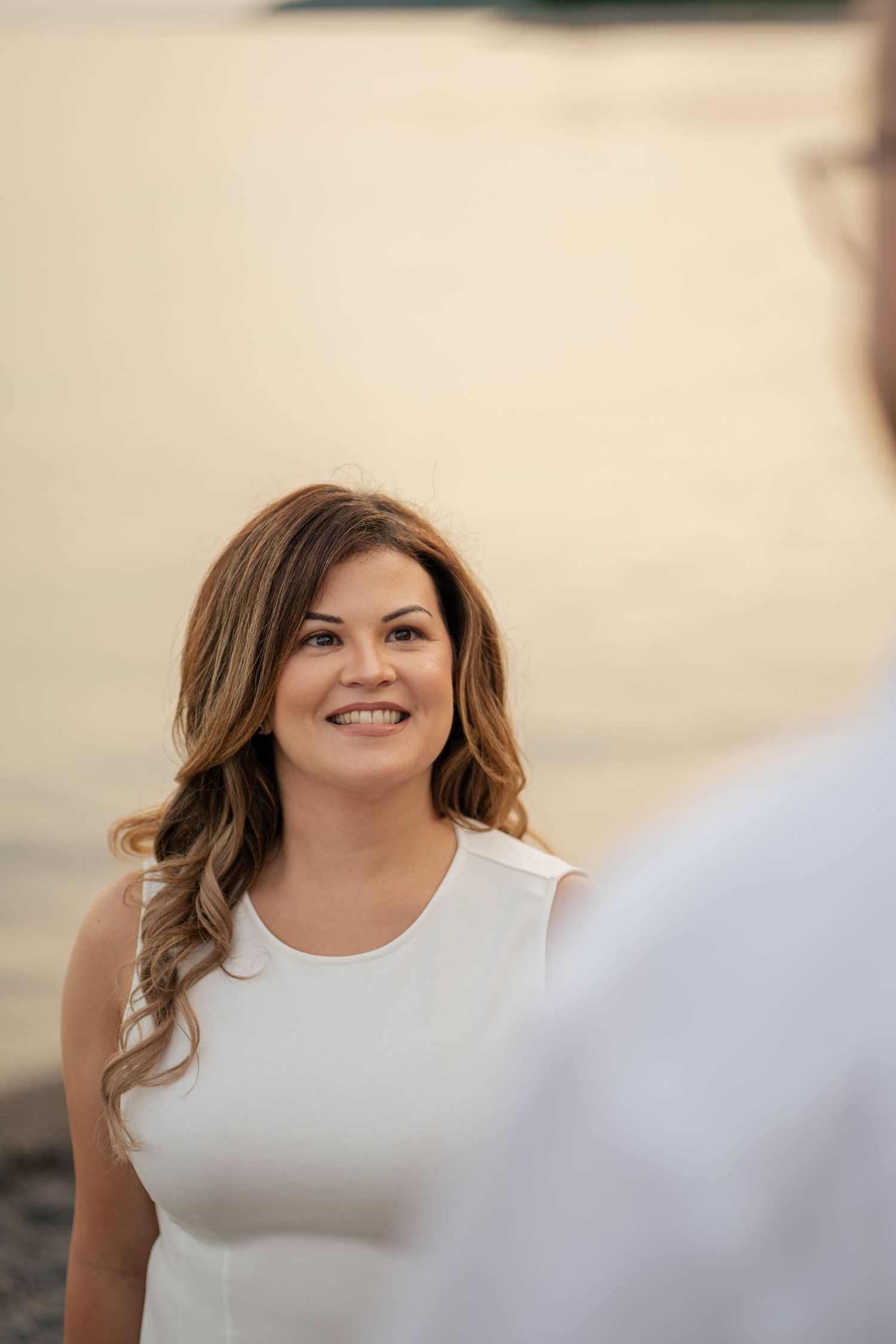 Close-up portrait of a smiling woman with wavy brown hair in a white top gazing at her partner by the water.