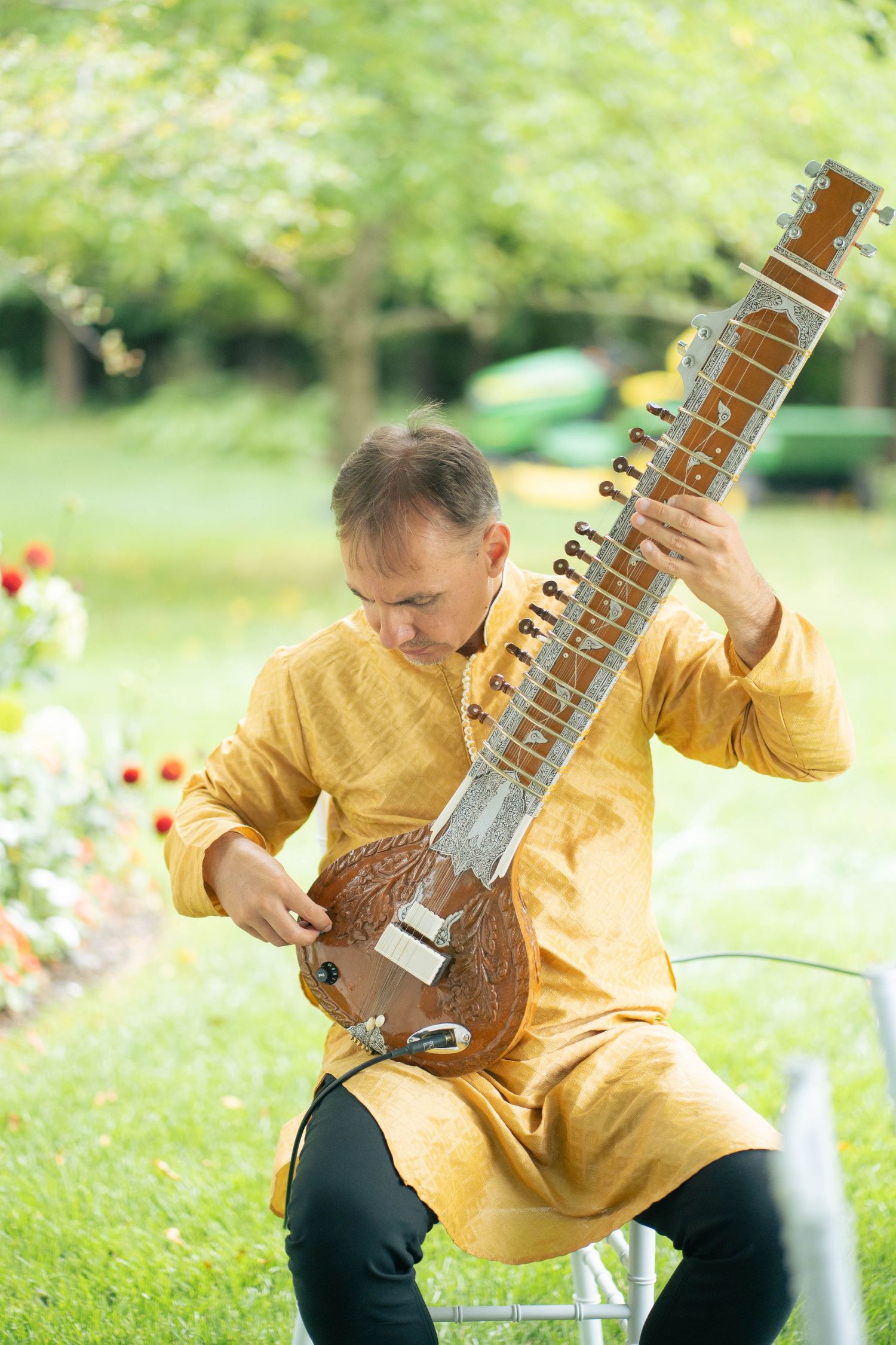 Man in a yellow kurta sits outdoors on a lawn playing a sitar, with greenery and flowers in the background.