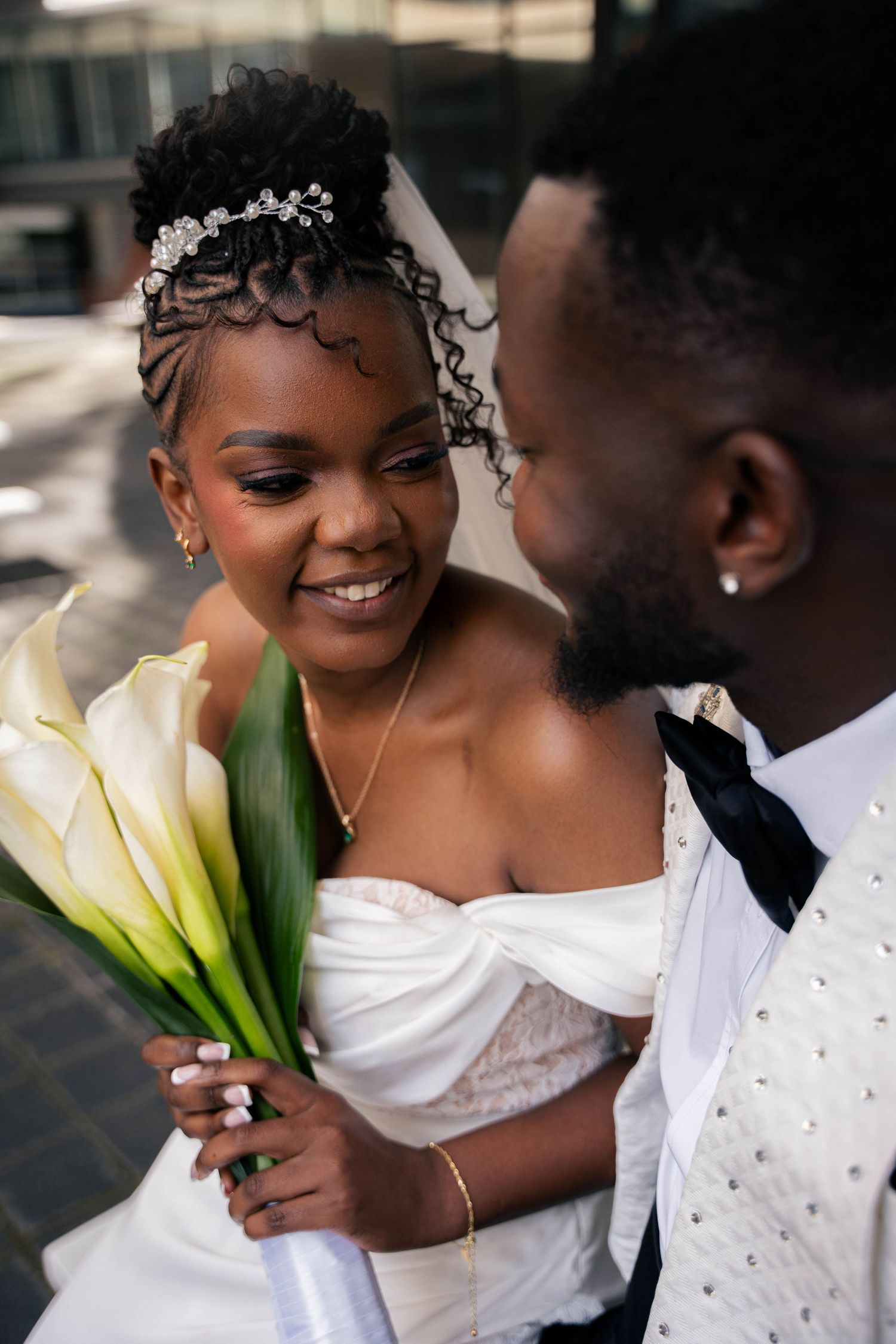 Joyful bride with a braided updo and jeweled hairpiece smiles beside her groom, holding a bouquet of white calla lilies.