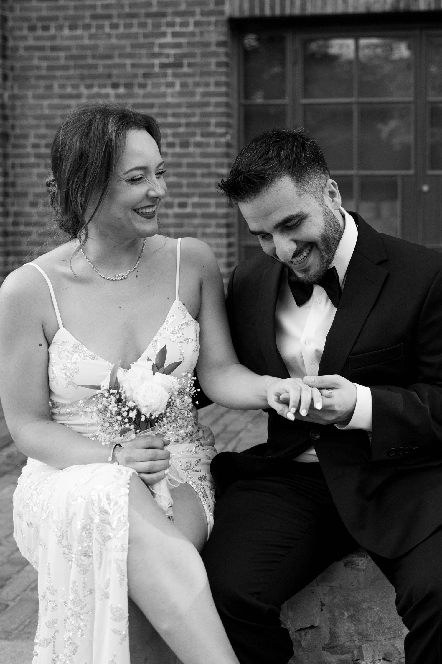 Smiling bride in a lace dress holding a bouquet looks down as her groom in a black tuxedo admires her ring in black and white.
