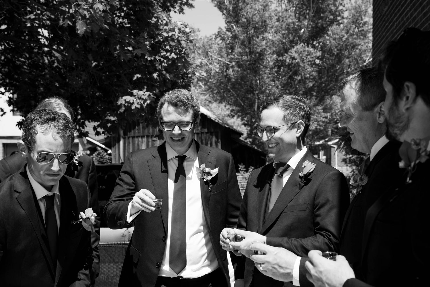 Groomsmen in suits laugh and chat outdoors as they hold small drinks before a wedding ceremony.
