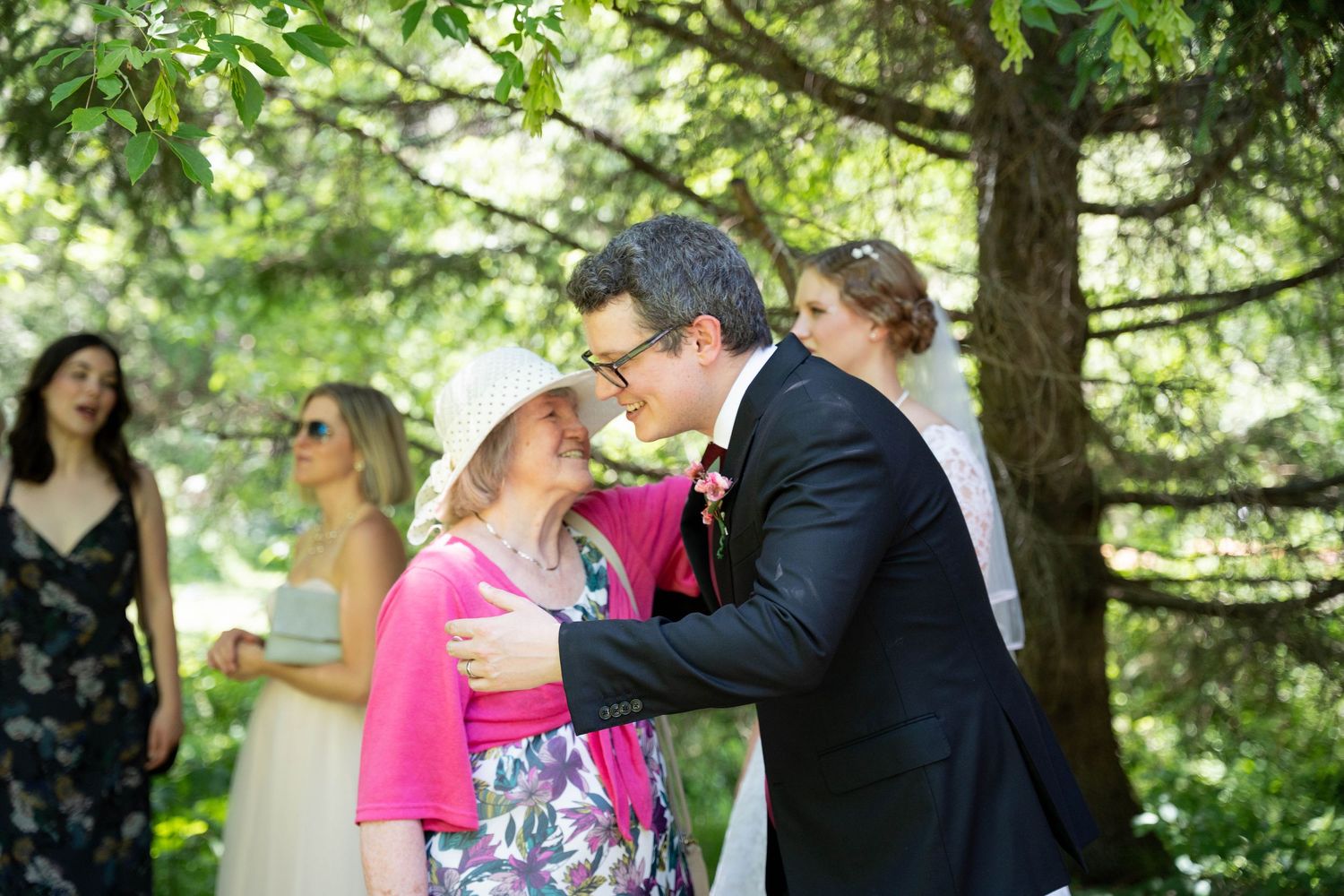 A groom in a dark suit hugs a smiling woman in a pink top and sun hat while the bride and guests stand nearby outdoors.