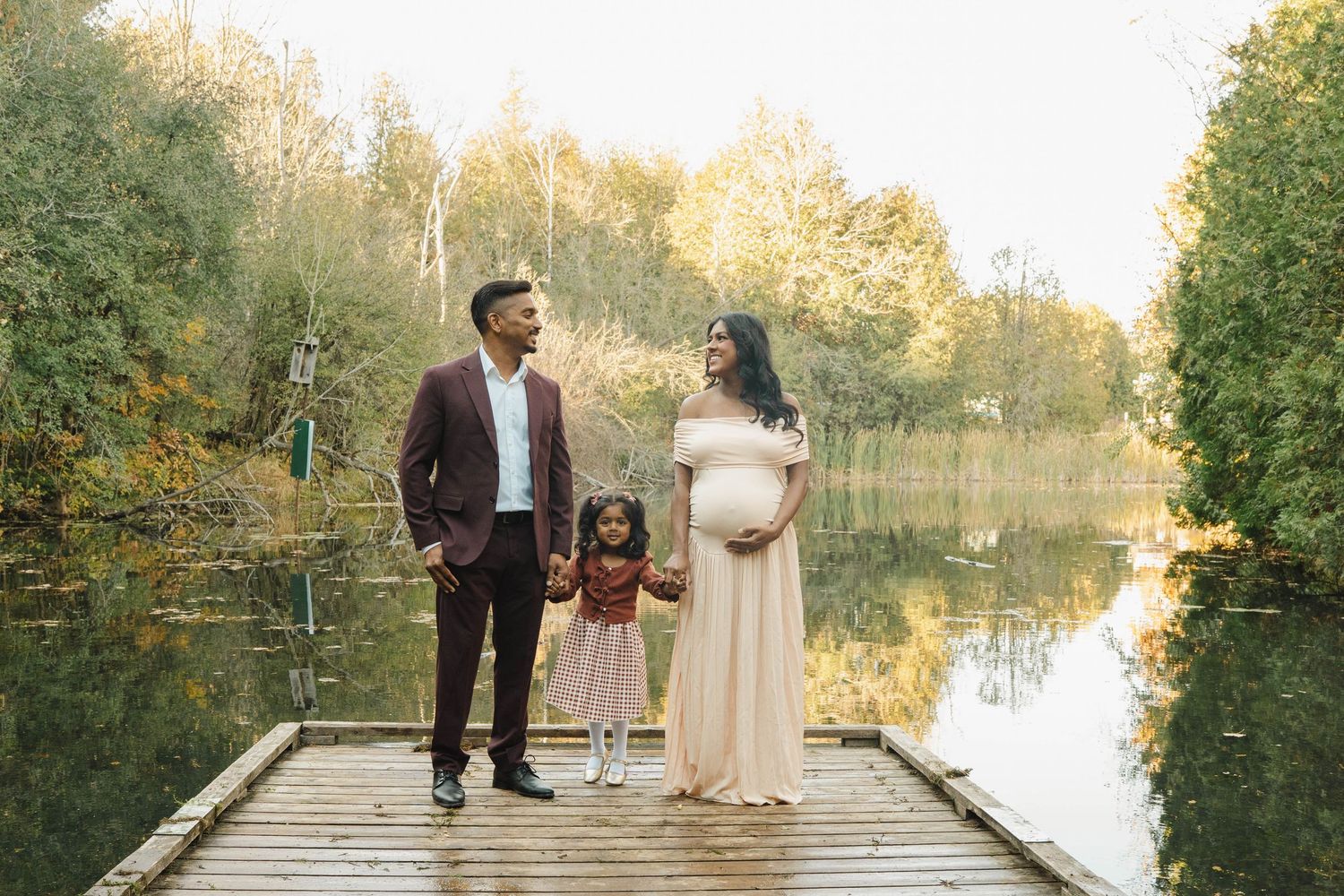 Expecting couple in formal attire holding hands with their young daughter on a wooden dock beside a calm autumn lake.