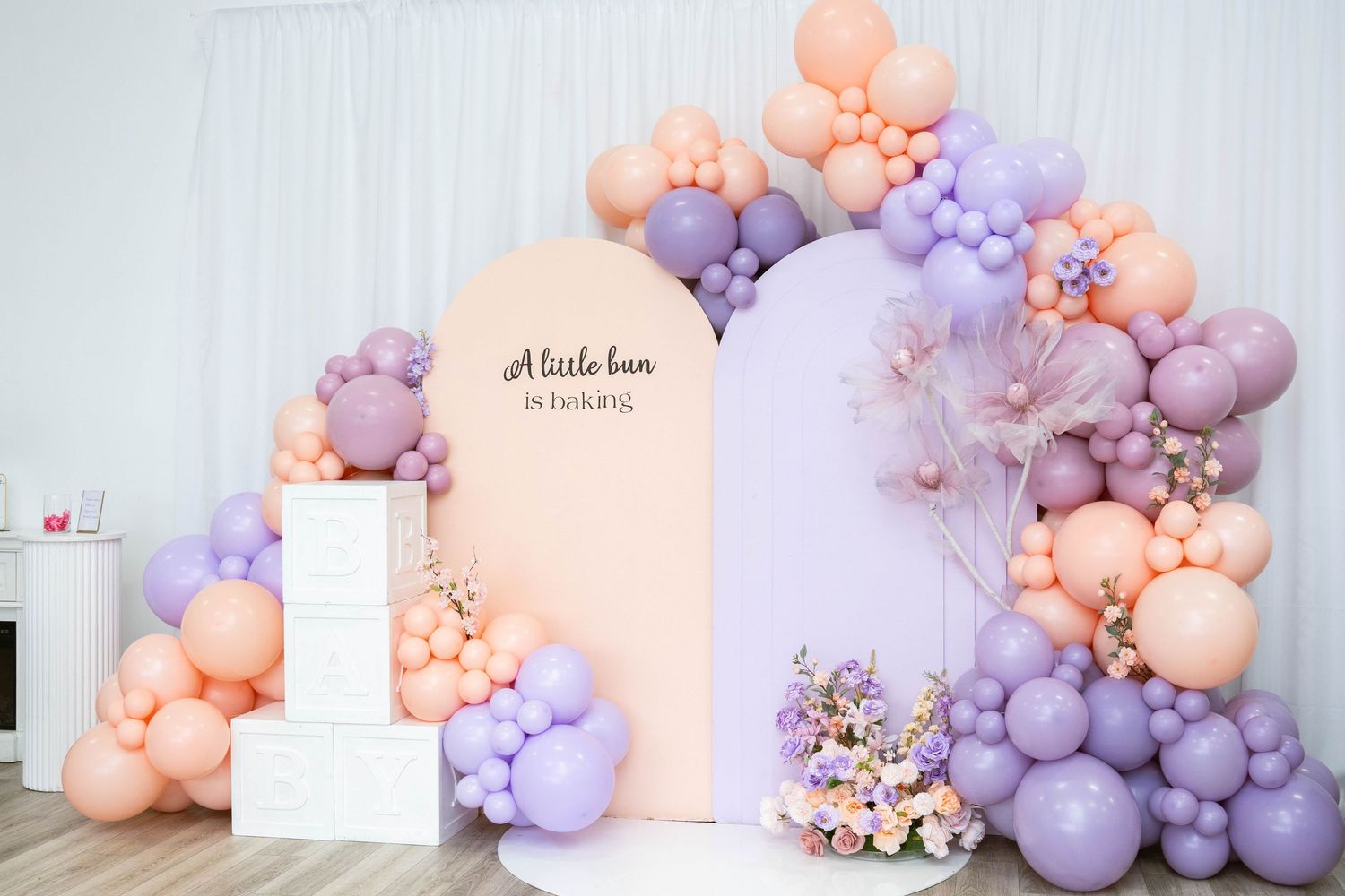 Pastel peach and lavender balloon garland arch with a sign reading A little bun is baking, beside white blocks and a floral arrangement.