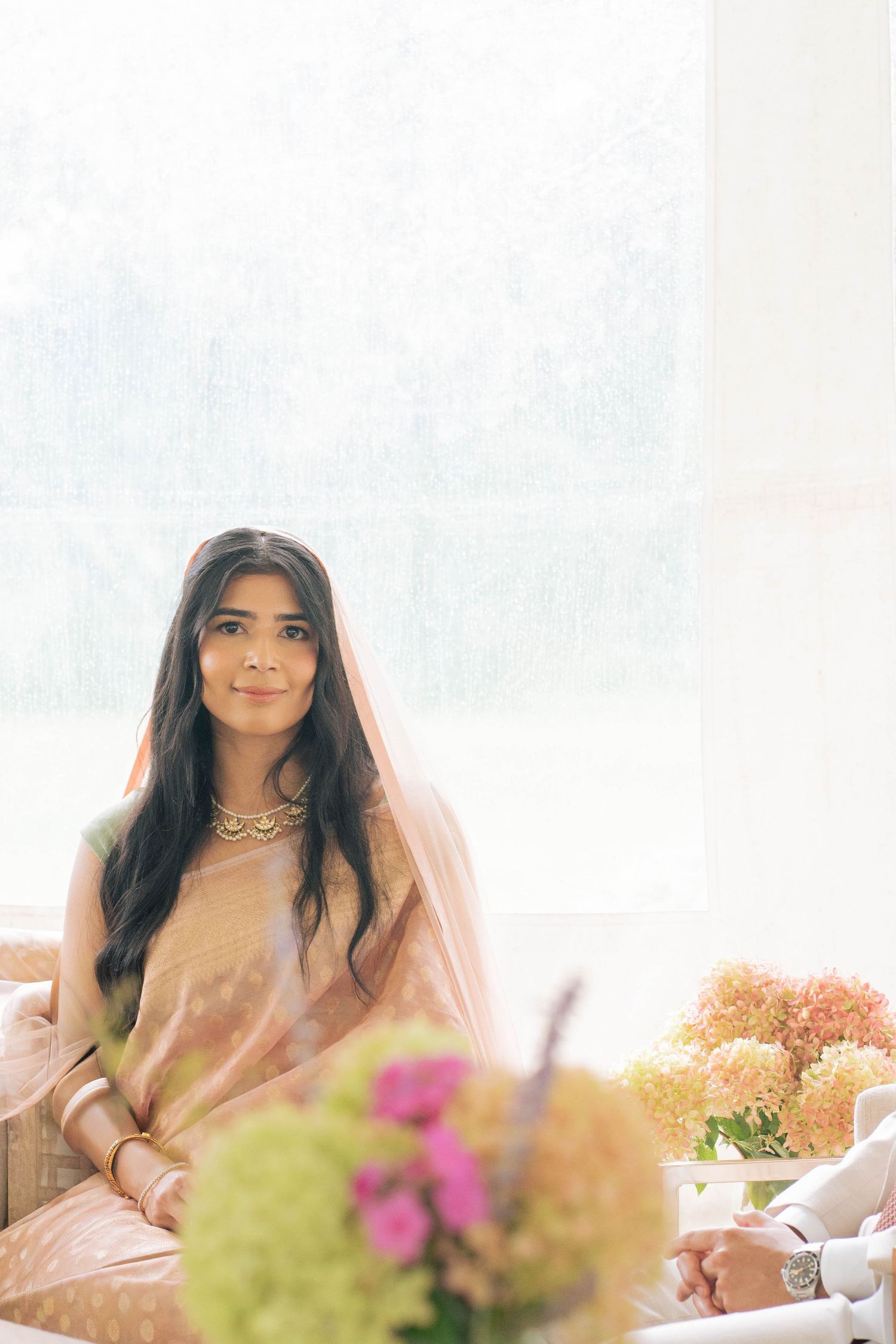 A serene bride in a peach sari, gold necklace and sheer veil sits by a bright window with pink and green flowers in the foreground.