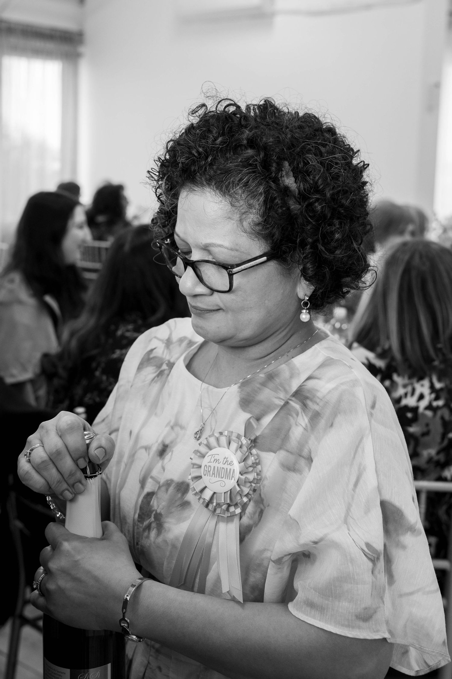A black and white portrait of a curly-haired woman wearing a grandma ribbon pin as she opens a champagne bottle at a party.