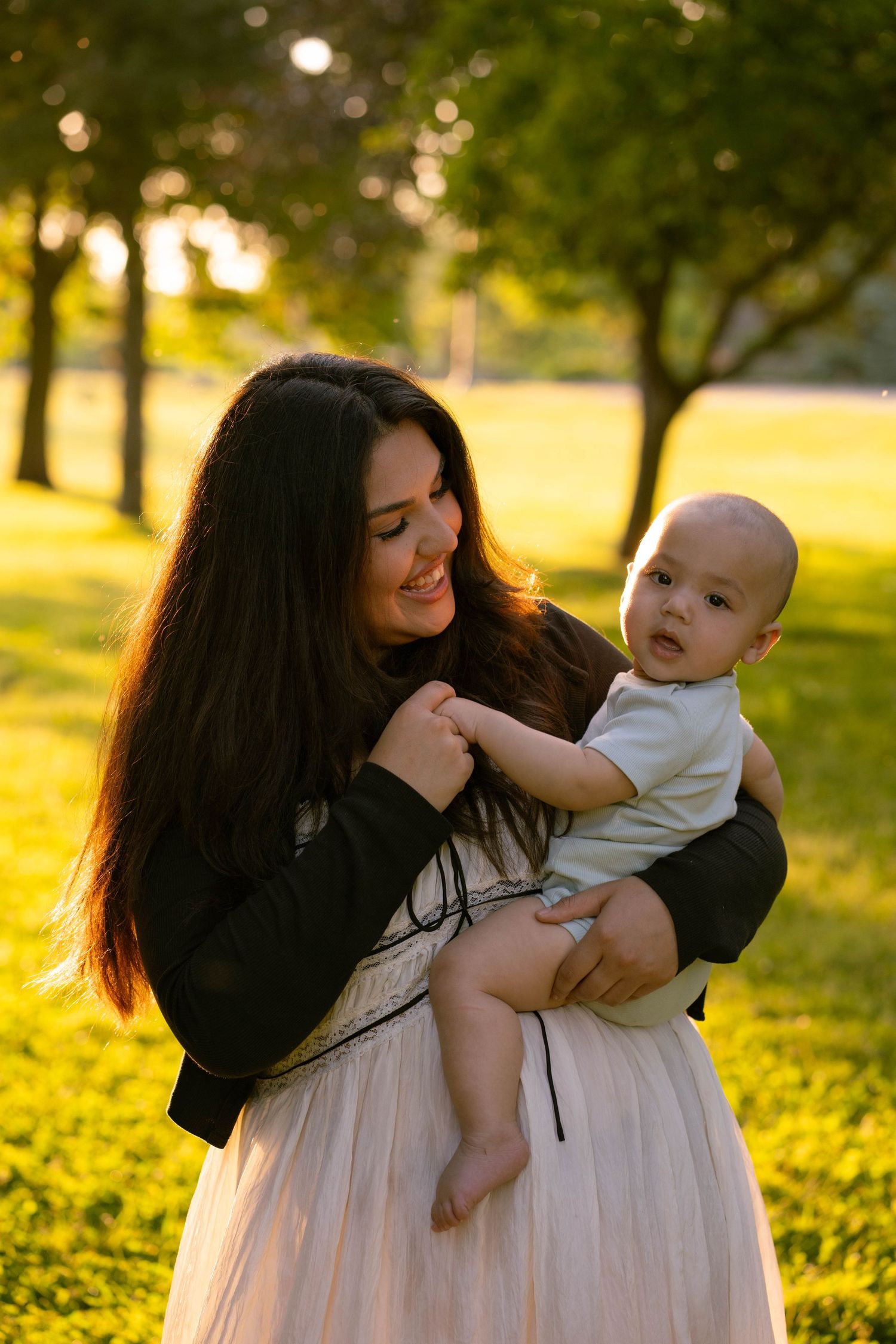 A smiling mother with long dark hair holds her baby in a sunlit park with golden evening light glowing behind them.