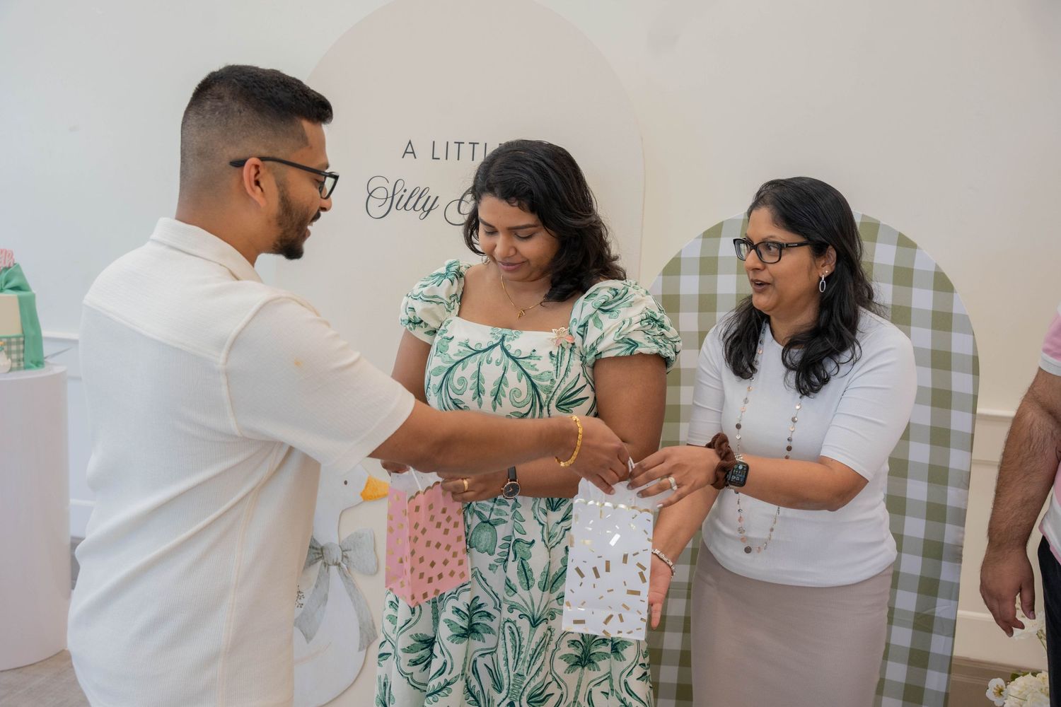 Man and women exchange small gift bags at a baby shower in front of a Little Silly Goose sign and green gingham backdrop.
