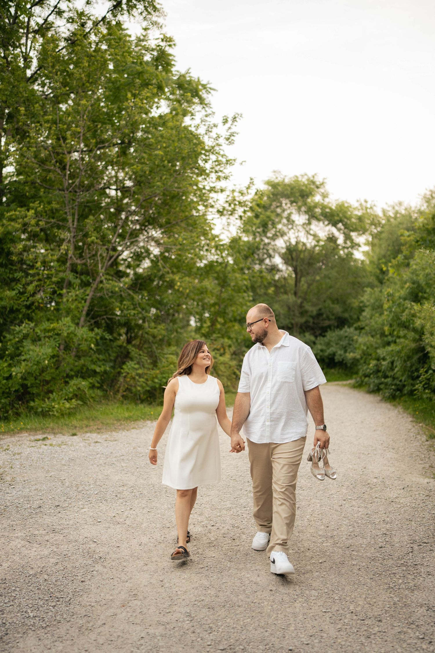 Couple walking hand in hand down a gravel trail surrounded by green trees, with the man carrying her silver heels.