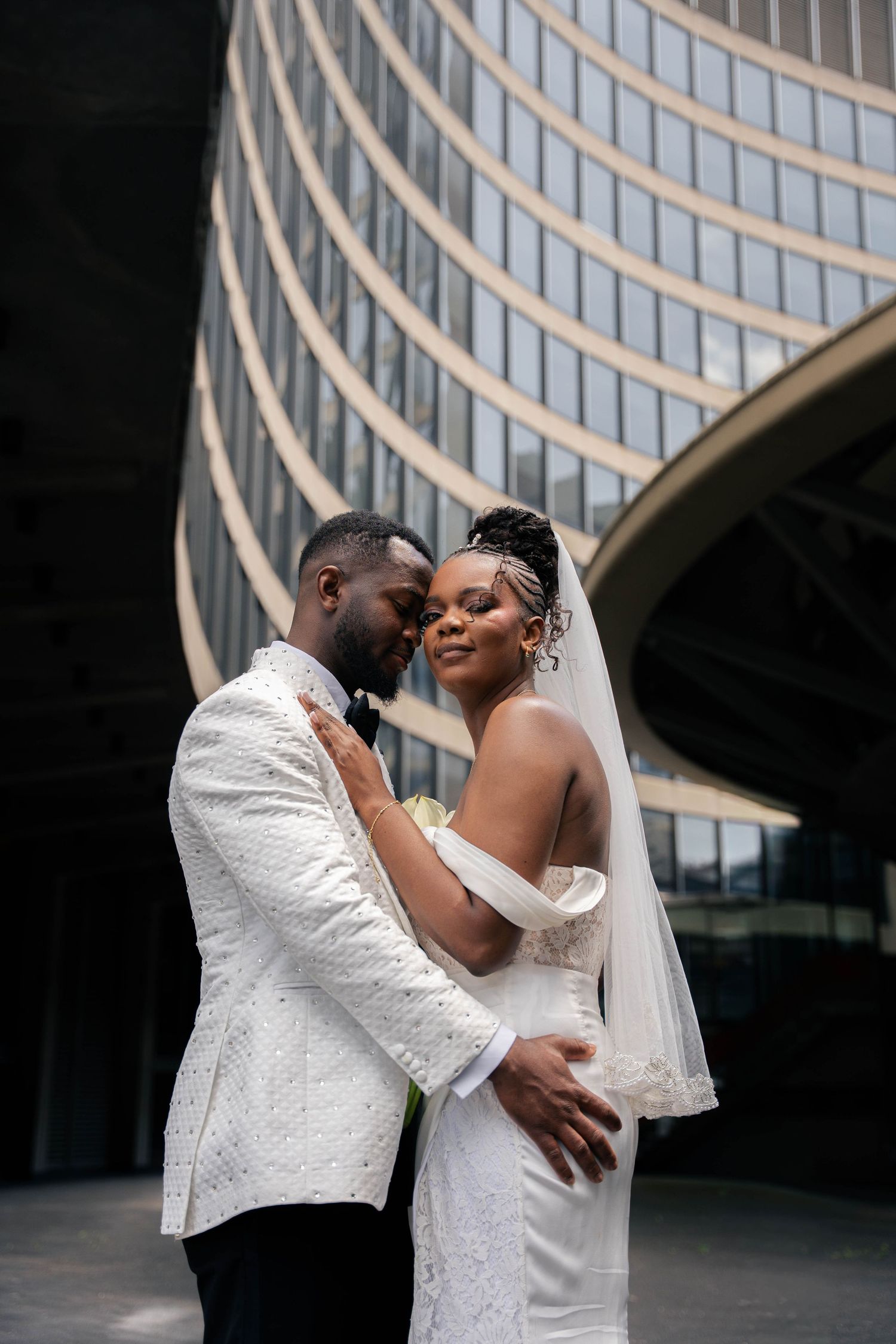 Groom in a white textured tuxedo embraces his bride in an off-shoulder gown and veil in front of a curved modern glass building.