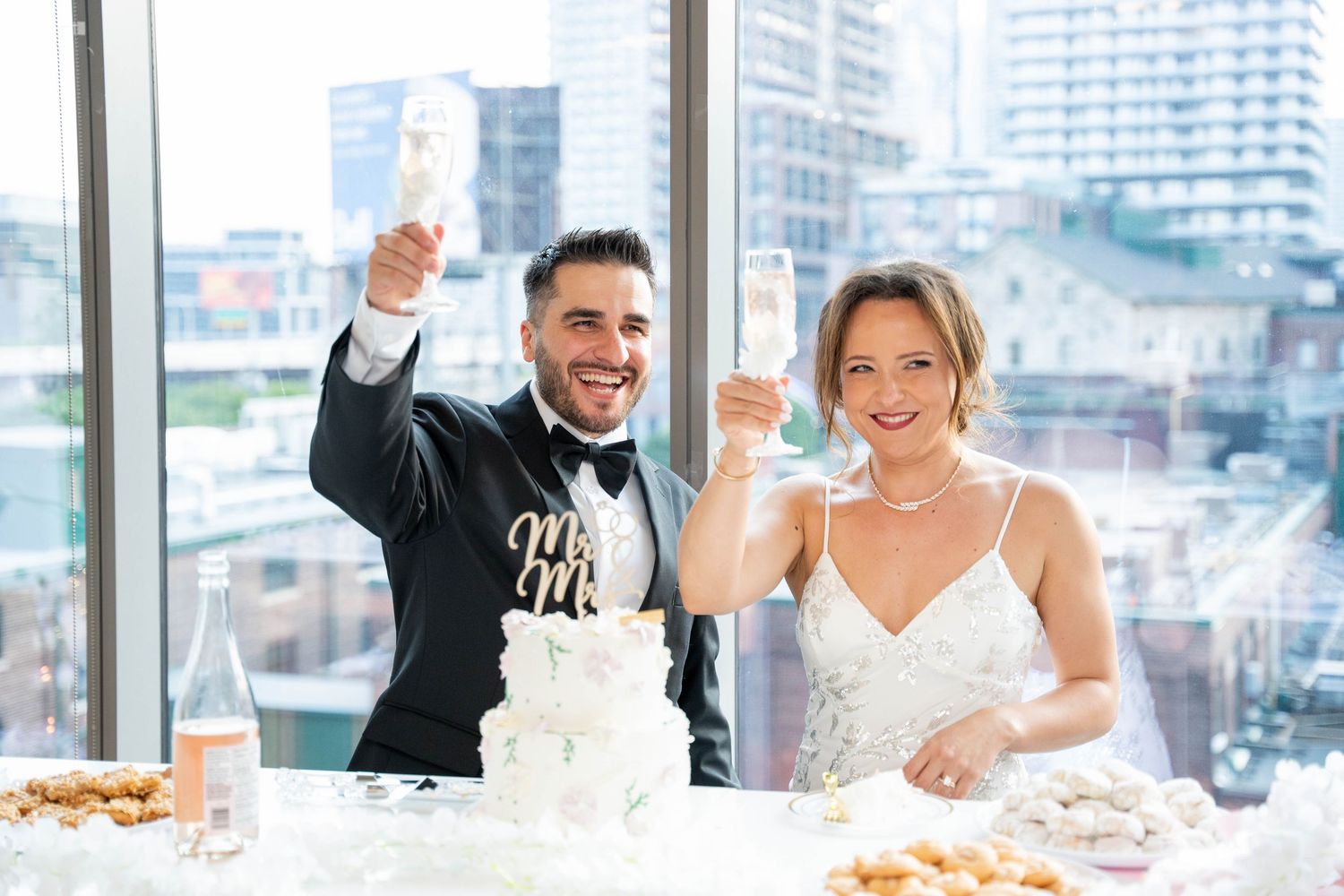 A smiling bride and groom raise champagne flutes in a toast beside their Mr and Mrs wedding cake with a city skyline behind.