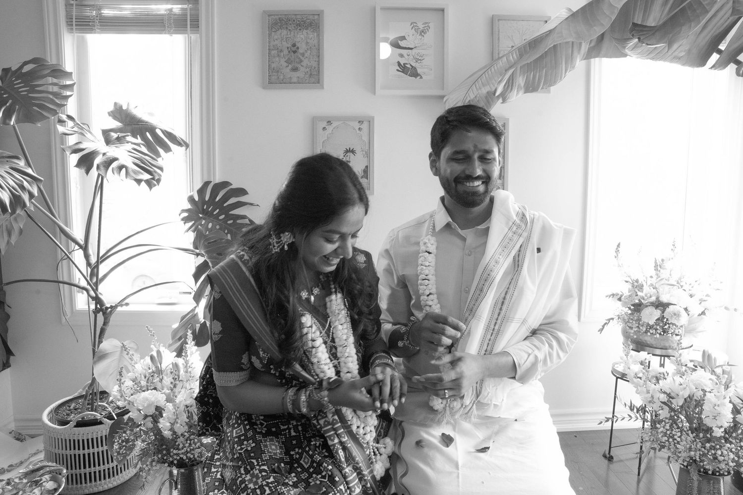 Black and white photo of a happy couple in traditional Indian wedding attire holding hands in a sunlit room with plants and flowers.