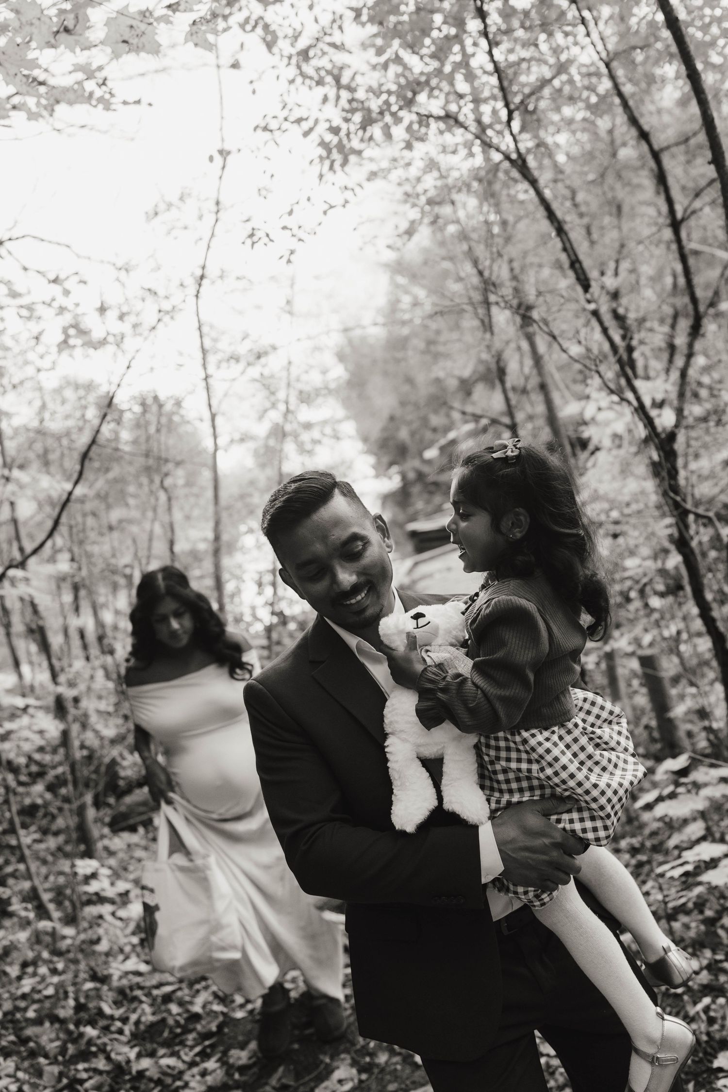Black and white maternity photo of a father carrying his daughter and a stuffed lamb on a wooded trail as the pregnant mother follows.