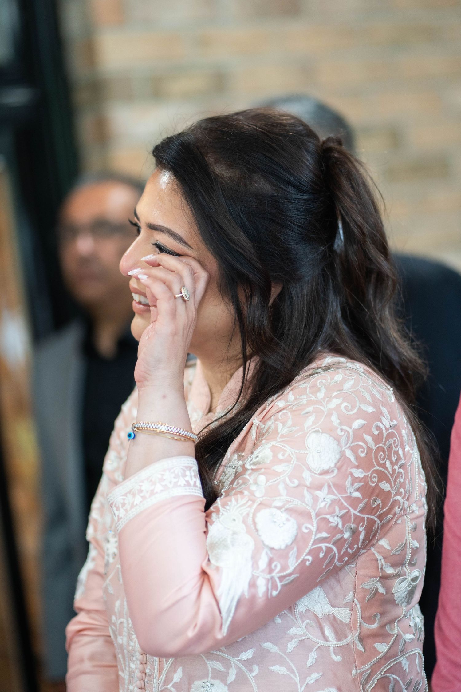A woman in an embroidered pink outfit wipes happy tears from her eye while guests look on behind her.