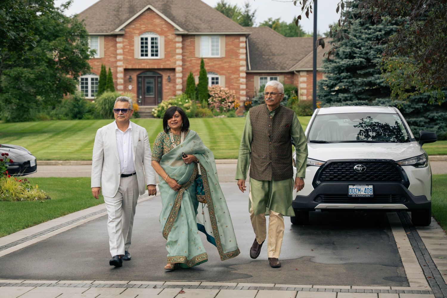 Three guests in elegant attire walk up a paved driveway toward a large brick home with a white SUV parked nearby.