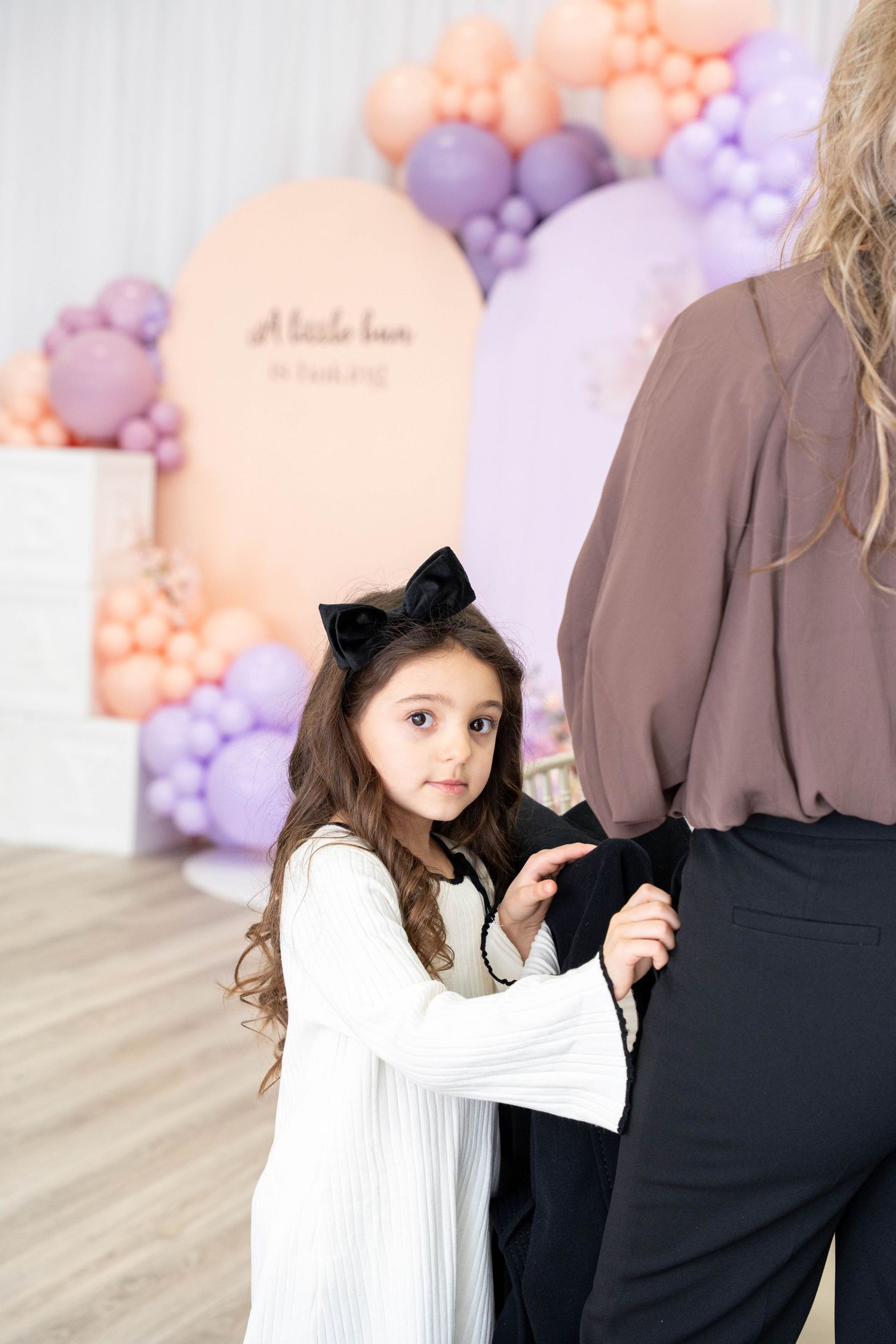 A young girl with a black bow tugs at an adult's sleeve in front of pastel balloons at a baby shower.