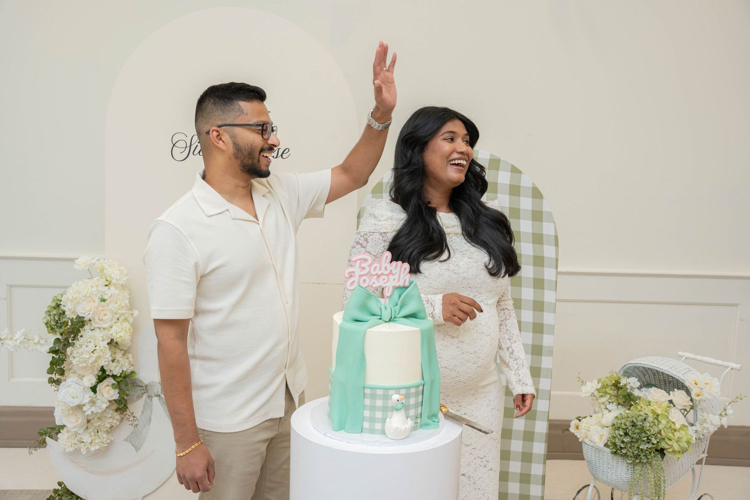 Man in glasses gestures excitedly as a smiling pregnant woman in a white lace dress stands beside a baby shower cake.