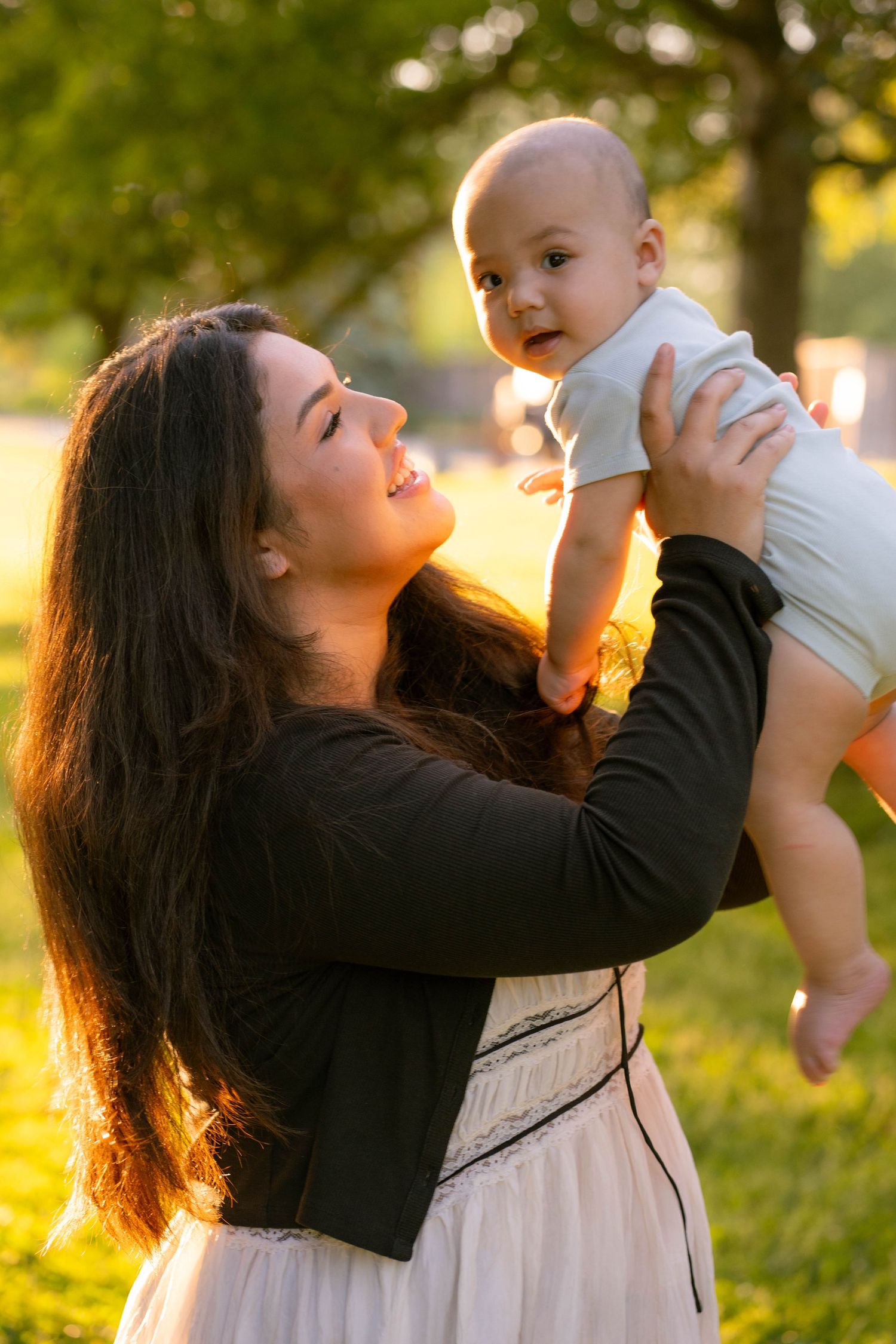 A laughing mother lifts her happy baby into the air in a glowing sunlit park during a family photo session.