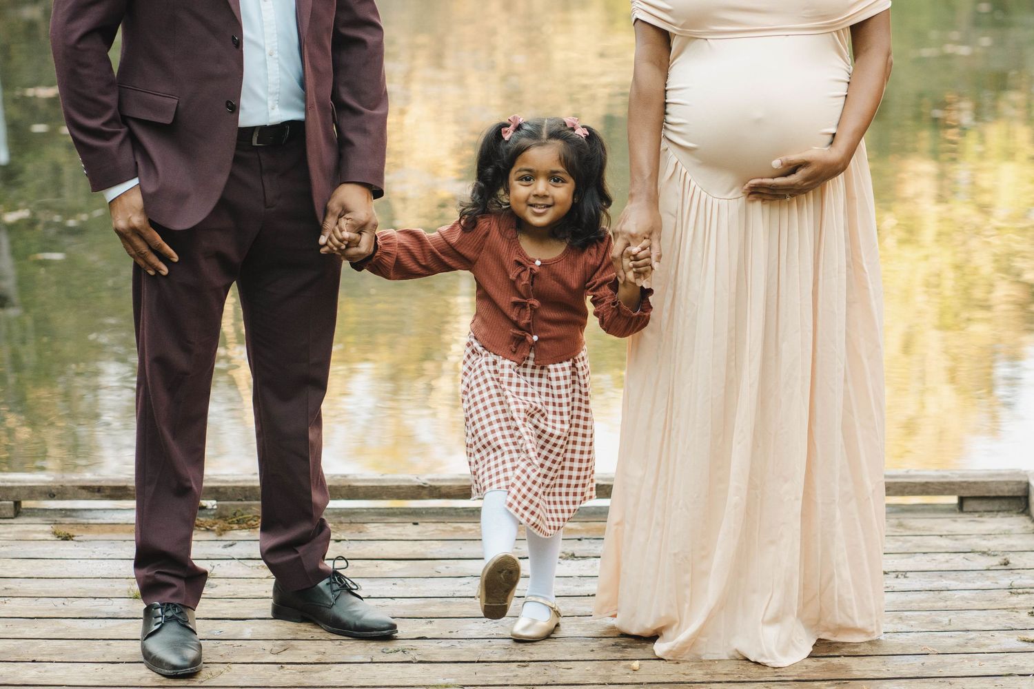 Pregnant mother in a peach gown and father in a suit hold hands with their little girl on a wooden dock beside a lake.