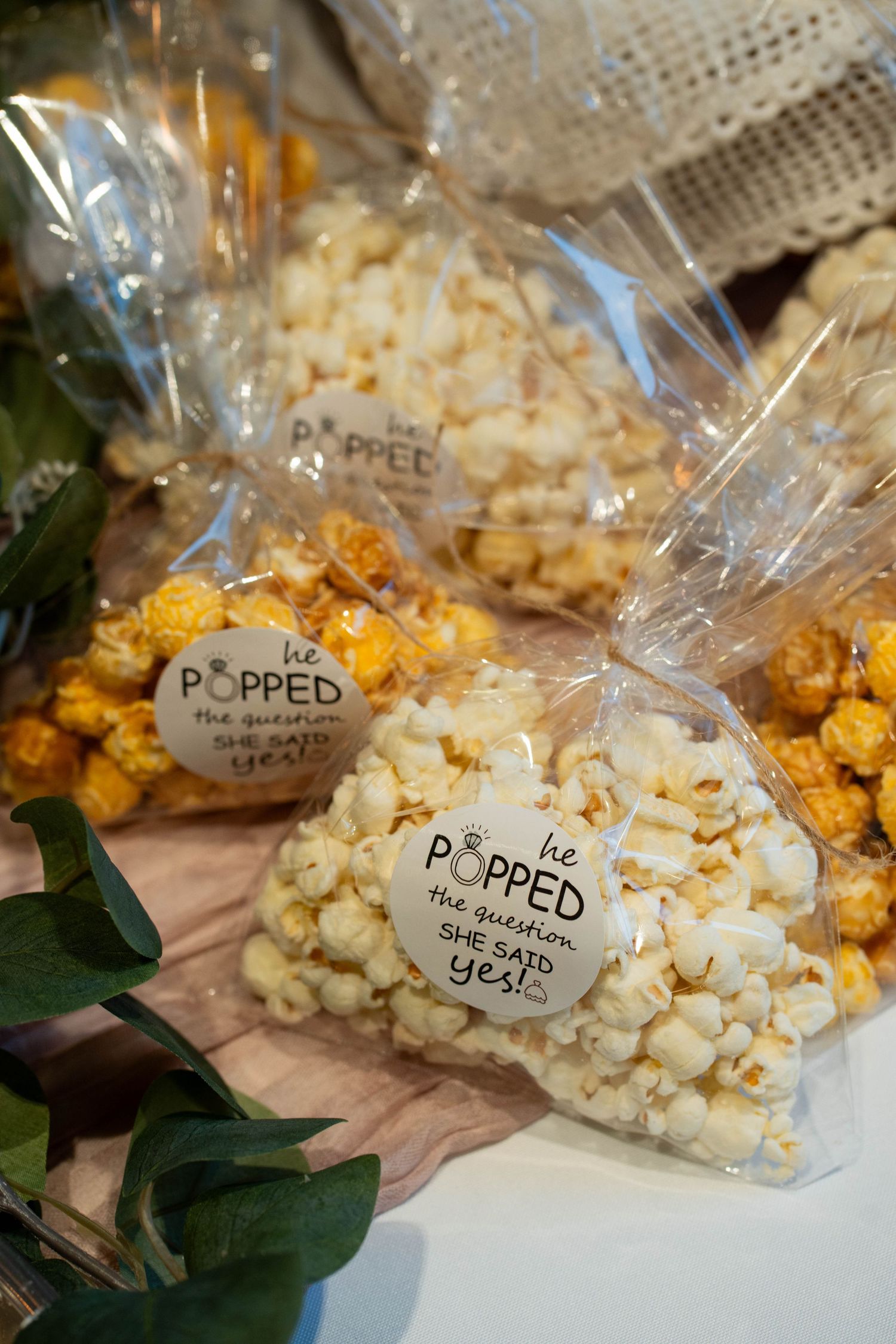 Bags of popcorn tied with tags that read He popped the question, she said yes, displayed on a pink table runner with greenery.