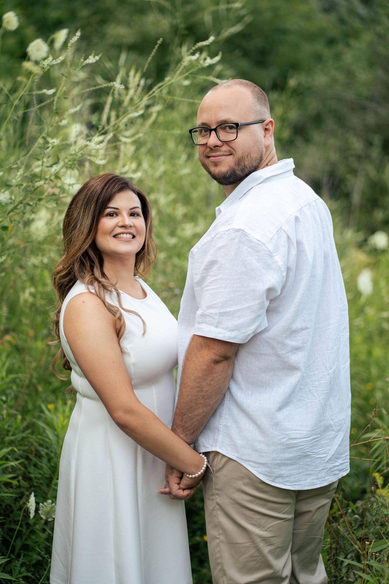 Happy engaged couple holding hands and smiling at the camera in a lush green meadow during a summer photo shoot.