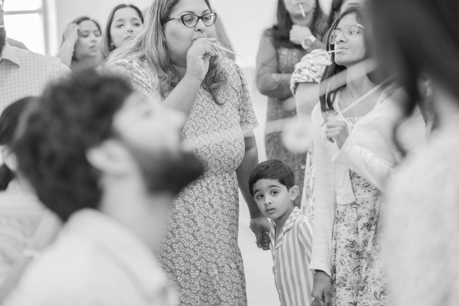 Black and white candid of baby shower guests singing and laughing while a young boy looks up from the crowd.