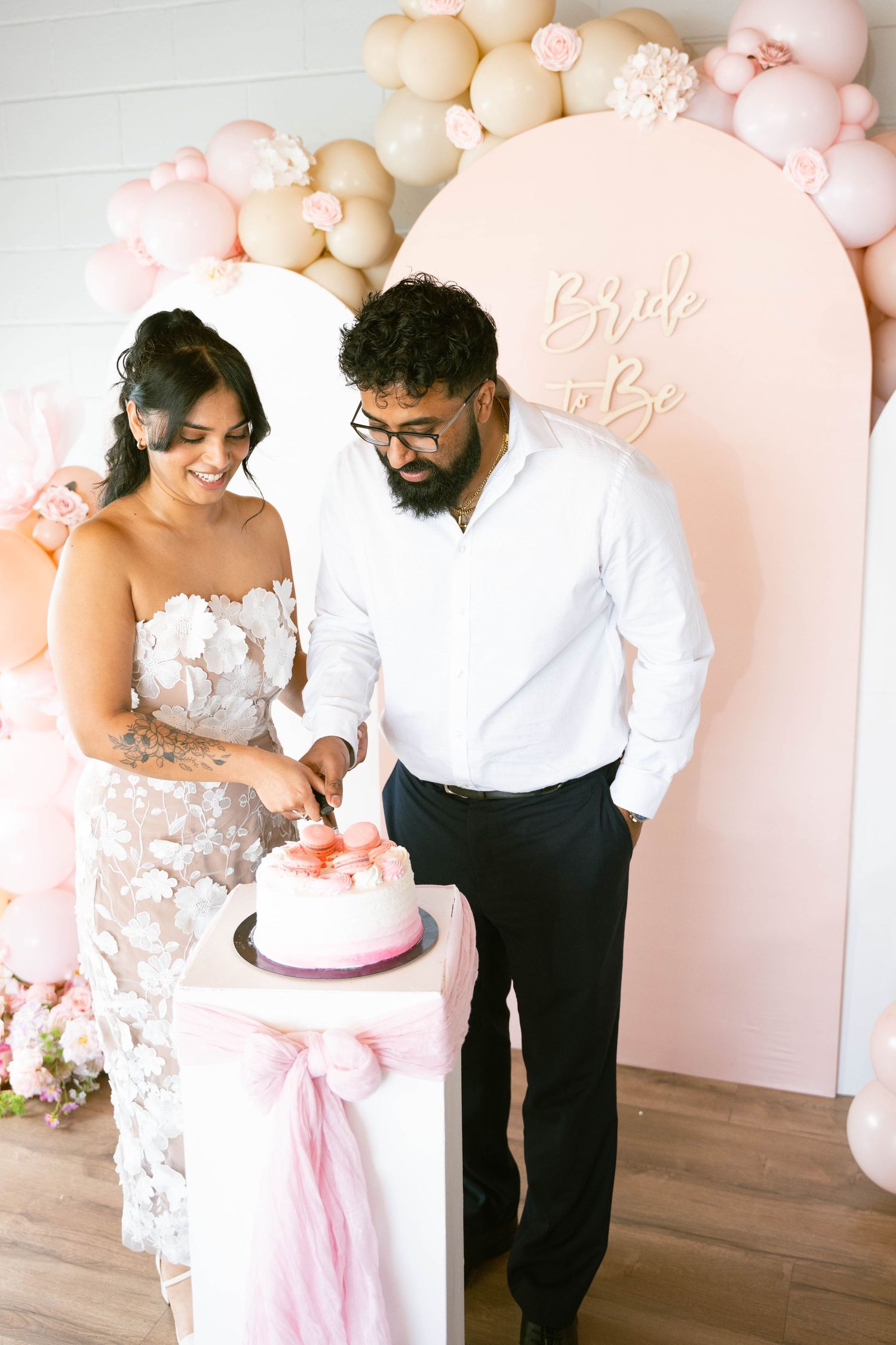 Couple cuts into a pink and white Bride to Be cake with macarons on a ribboned pedestal at their bridal shower.