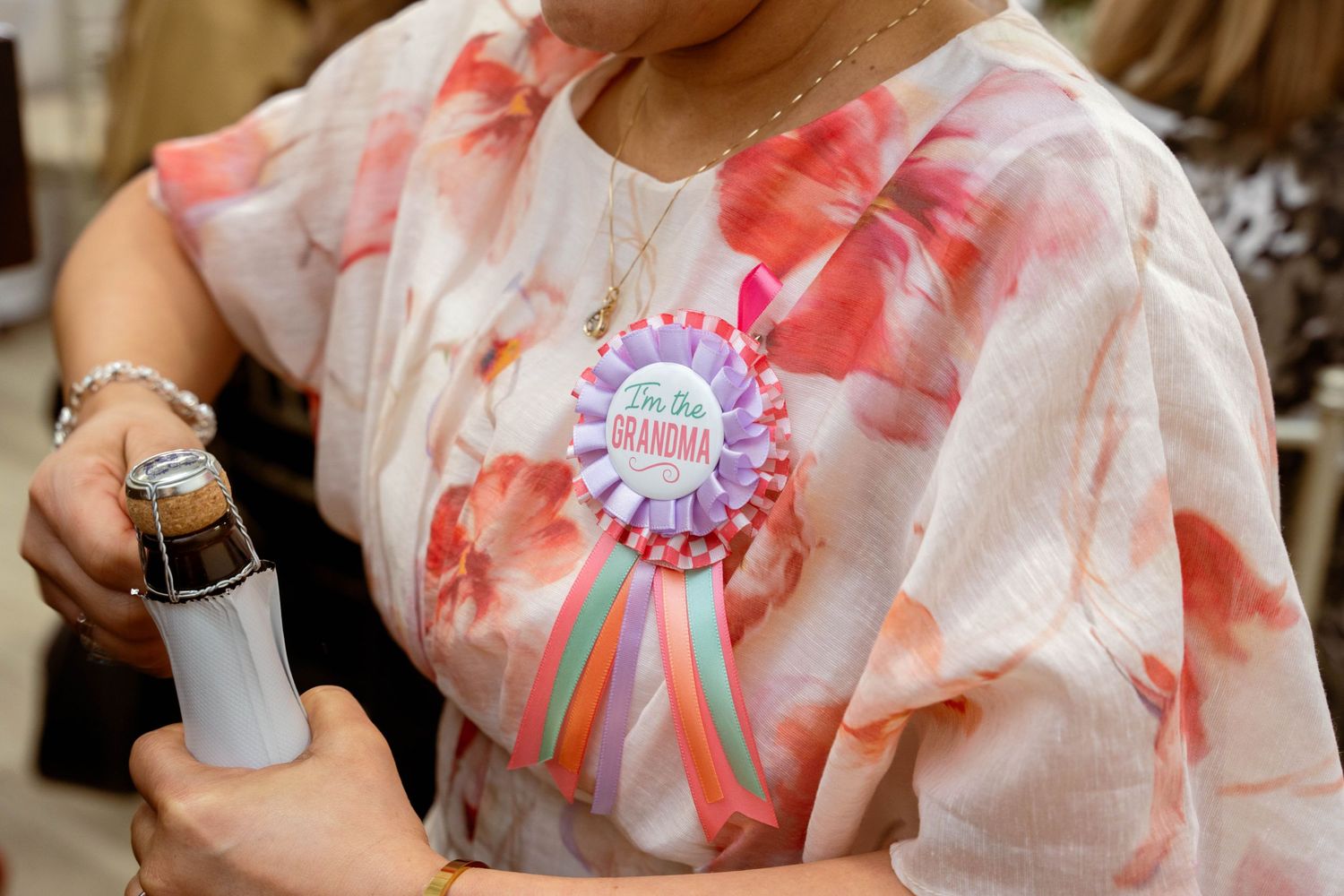 A woman in a peach floral blouse wears a grandma ribbon rosette while holding a champagne bottle at a baby shower.