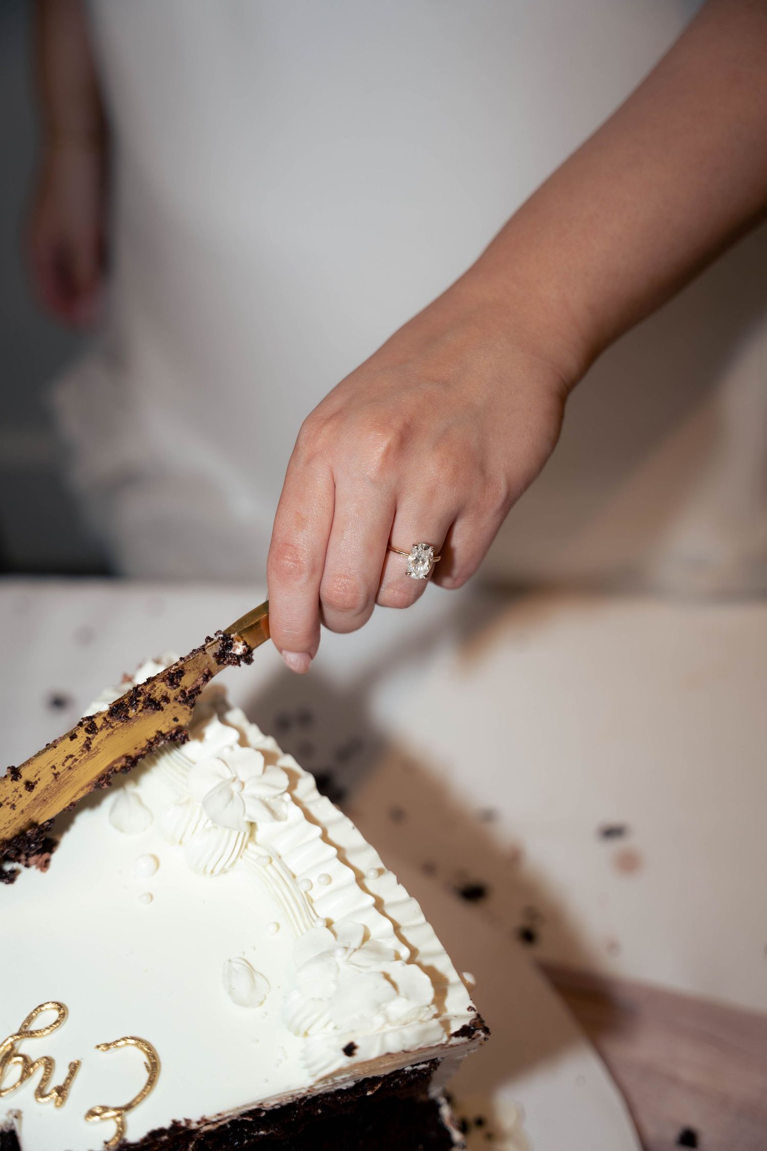 A hand wearing a diamond engagement ring slices a white frosted cake with gold lettering and chocolate layers using a knife.