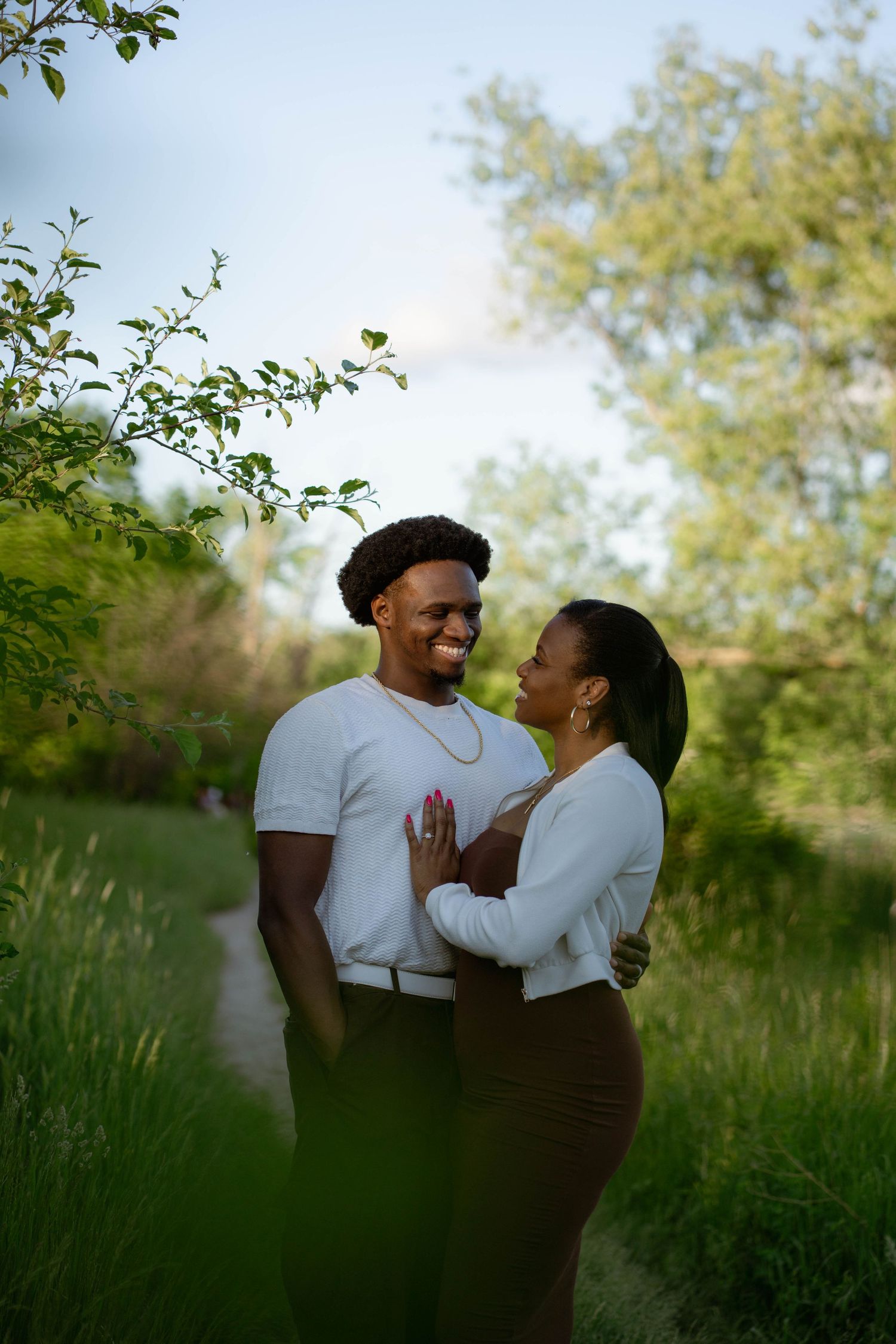An engaged couple embraces and smiles on a leafy park trail during an outdoor engagement photo session.