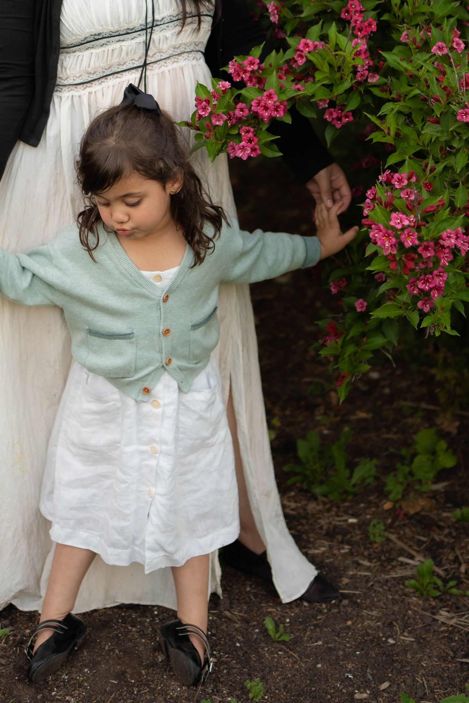 A little girl in a green cardigan and white dress reaches for her mother's hand beside a pink flowering bush.