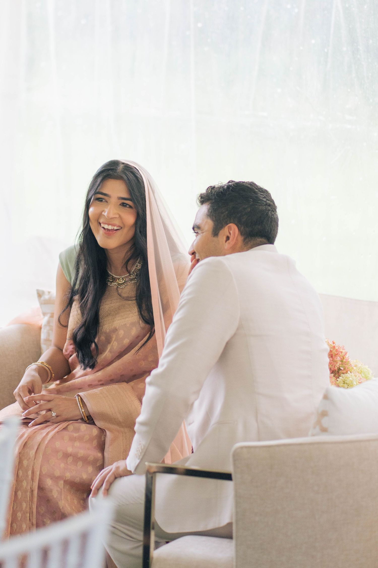A joyful bride in a peach sari and sheer veil laughs warmly beside her groom, who wears a cream suit, during an intimate wedding ceremony.